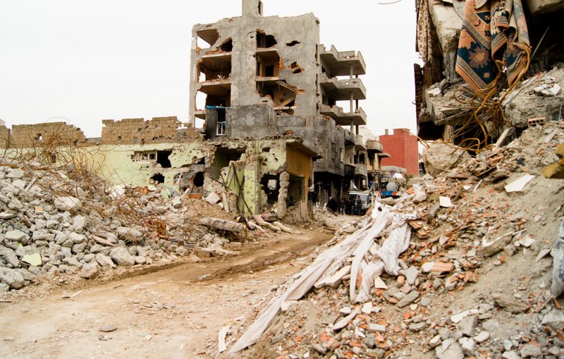 L'image montre un paysage urbain dévasté par la guerre ou une catastrophe. On peut voir des bâtiments partiellement détruits, présentant des murs troués et des structures effondrées. Des débris encombrent le sol, et des morceaux de béton et des matériaux de construction sont éparpillés partout. La rue semble désolée, soulignant l'impact de la destruction sur l'environnement urbain. Les couleurs sont sombres et tristes, renforçant le sentiment de désespoir et de désolation.