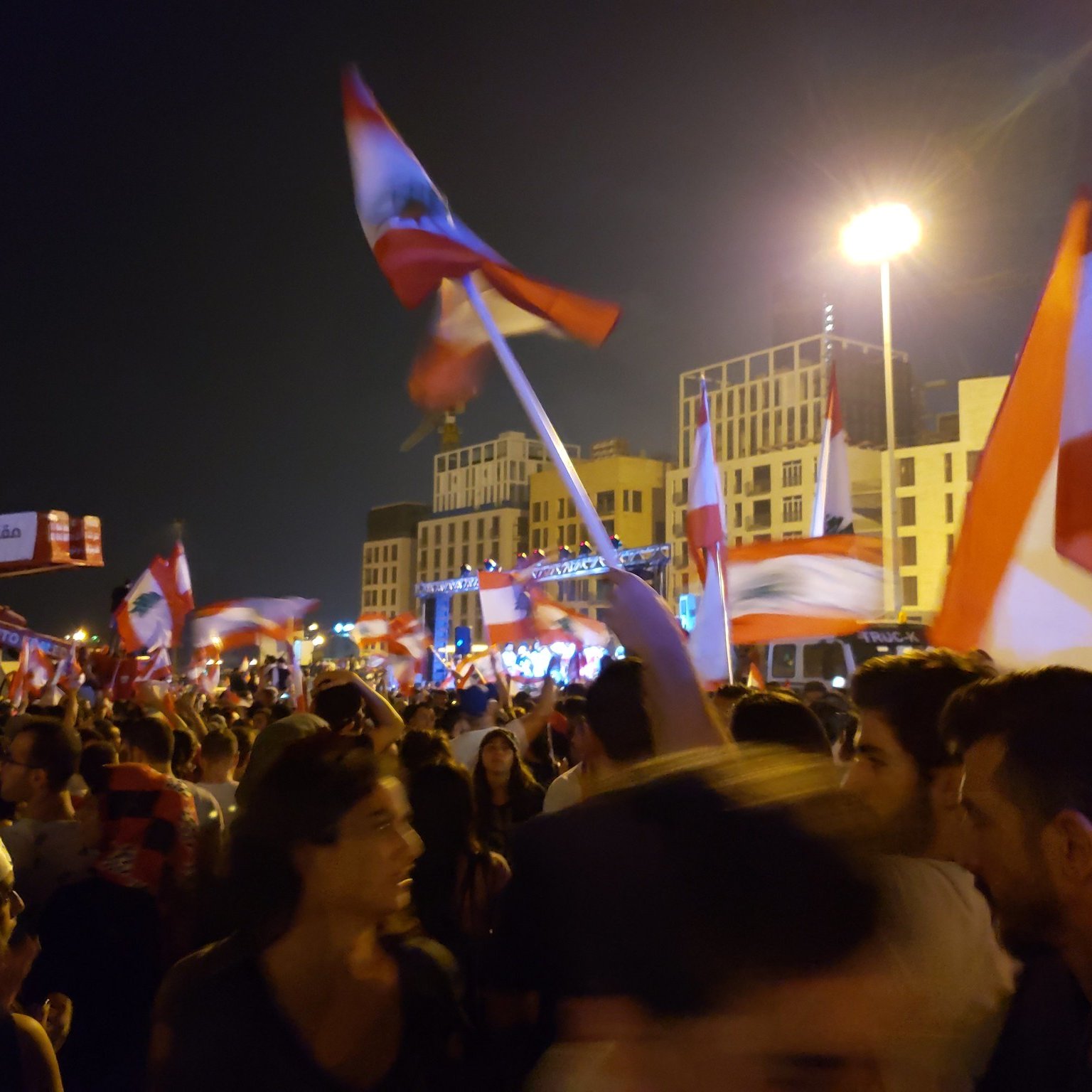 L'image montre une foule rassemblée à la nuit tombée, tenant des drapeaux libanais. Les gens semblent être en plein mouvement, probablement en train de célébrer ou de manifester. On aperçoit une scène à l'arrière-plan, éclairée par des lumières, ce qui suggère un événement festif ou politique. L'atmosphère est dynamique, remplie d'énergie, avec des expressions de joie et d'engagement parmi les participants.