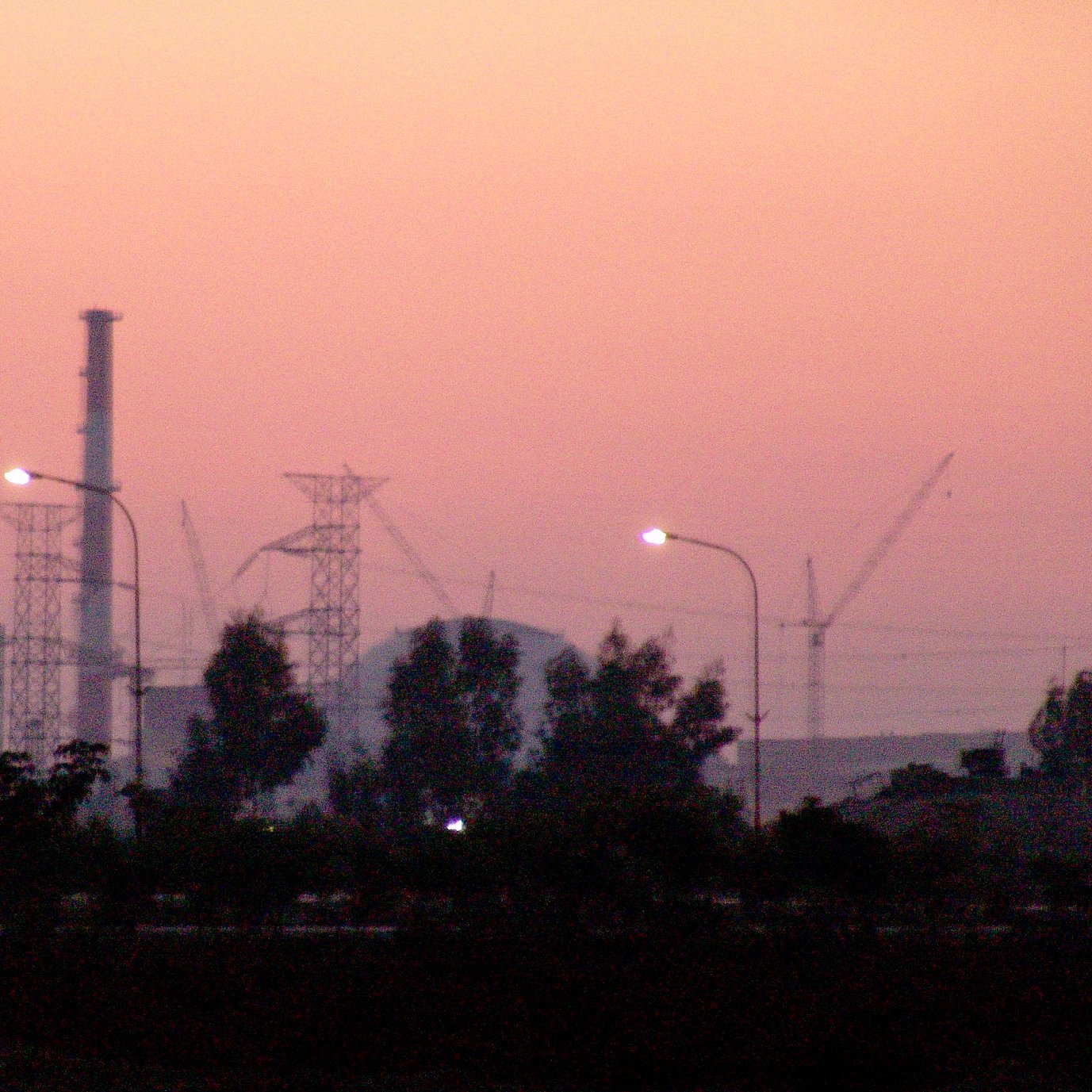 L'image montre une scène de crépuscule avec un ciel teinté de rose et d'orange. Au premier plan, on peut voir des arbres ainsi que des lampadaires allumés. En arrière-plan, se dressent des structures industrielles, notamment des cheminées et des pylônes électriques, ainsi que des grues de construction, ce qui suggère une activité industrielle en cours. L'atmosphère est calme et paisible, malgré la présence des installations.