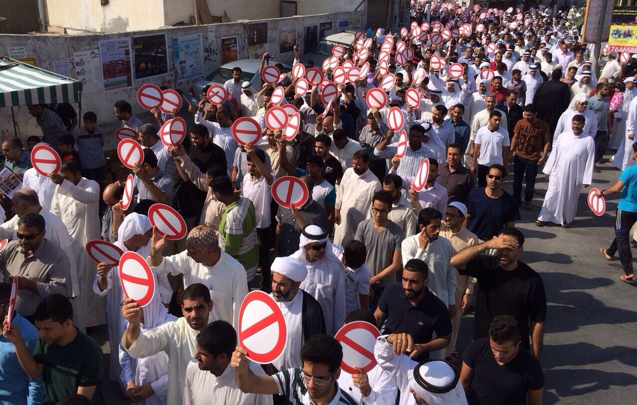 L'image montre une grande foule de personnes rassemblées dans une rue. Les manifestants brandissent des pancartes avec un symbole de cercle rouge barré, souvent utilisé pour signifier une interdiction. Les participants portent des vêtements variés, certains en tenue traditionnelle. On observe des hommes et des femmes de différentes origines, tous unis dans leur démarche. L'environnement urbain en arrière-plan, avec des bâtiments et des affiches, suggère un contexte de protestation ou de manifestation.