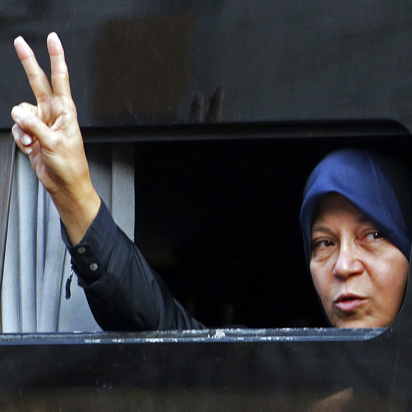 L'image montre une femme qui fait un signe de paix avec sa main, en levant deux doigts. Elle est visible à travers une fenêtre légèrement ouverte d'un véhicule. La femme porte un hijab. Son expression semble déterminée et engagée. Le fond est sombre, ce qui met en avant son geste et son visage.