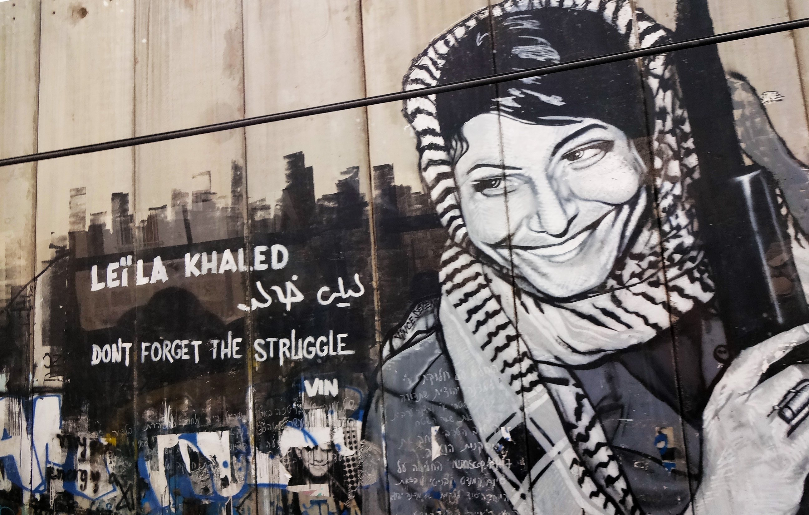 L'image présente une fresque murale représentant Leïla Khaled, une figure emblématique de la résistance palestinienne. Elle est dépeinte avec un sourire, portant une écharpe typique et tenant un fusil. À côté de son portrait, on peut lire des inscriptions en anglais et en arabe, telles que "Don't forget the struggle" et son nom. L'arrière-plan est constitué d'un mur, accentuant le message de lutte et de mémoire associé à son image. Des graffitis colorés et des marques diverses décorent également la surface, ajoutant à l'atmosphère militante et artistique du lieu.