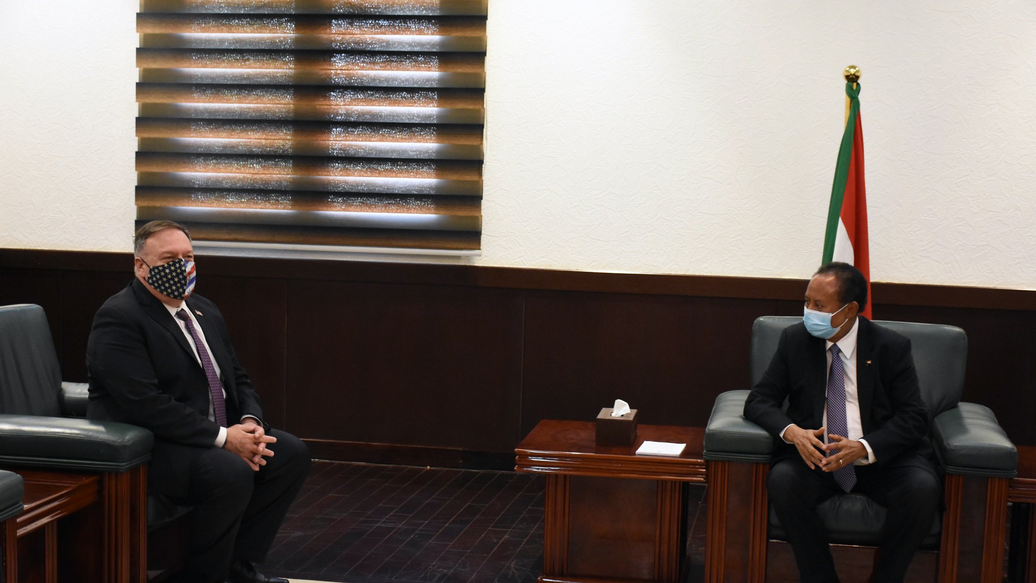 در این تصویر، دو مرد در یک اتاق رسمی نشستهاند. یکی از آنها که سمت چپ تصویر است، ماسک دارد و ظاهری رسمی با کت و شلوار پوشیده است. مرد دیگر در سمت راست نیز لباس رسمی به تن دارد و به طرف مرد اول نگاه میکند. در پسزمینه، پردههای پنجره و پرچم یک کشور مشخص است. فضا به وضوح رسمی و موقر به نظر میرسد و به احتمال زیاد مربوط به یک ملاقات دیپلماتیک است.