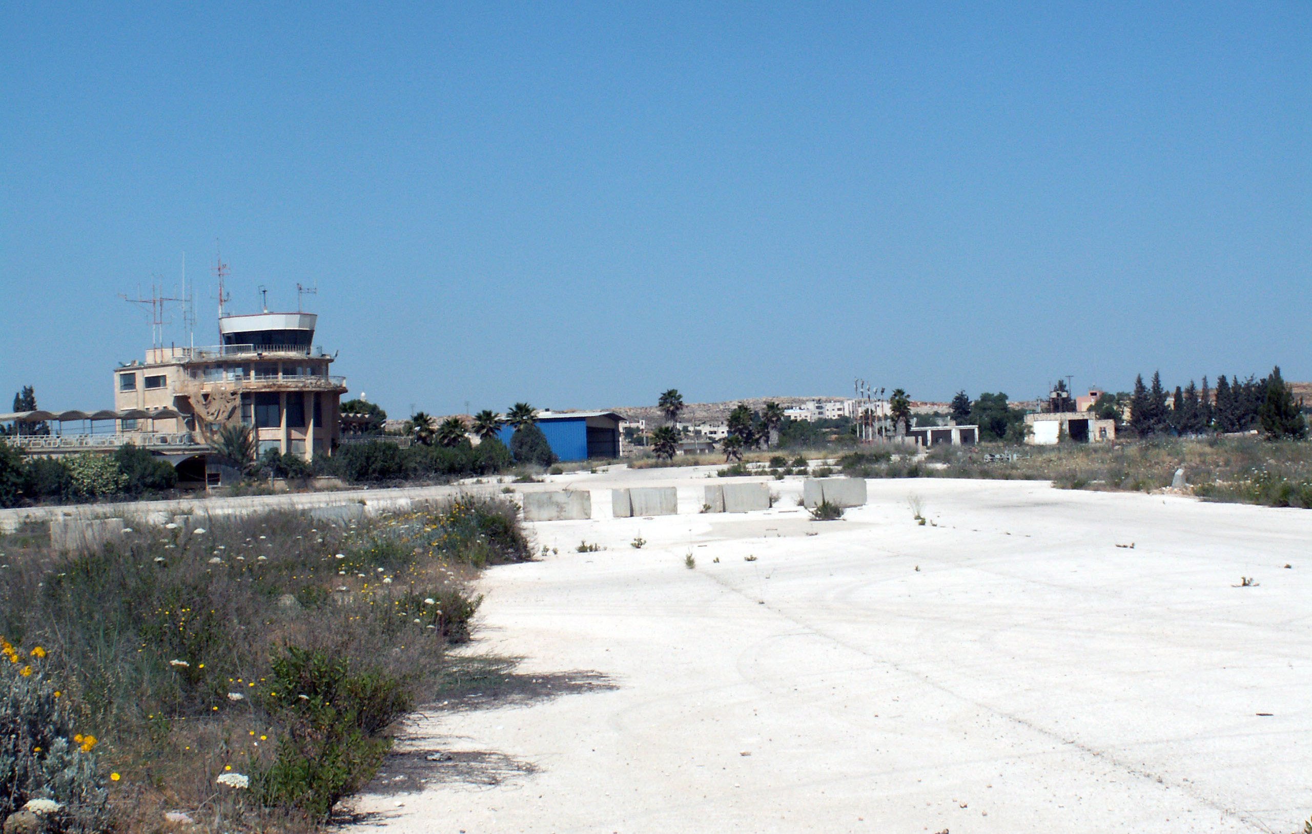 L'image montre un paysage de terrain vague, probablement une ancienne installation ou un aéroport désaffecté. Au loin, on peut voir un bâtiment de type tour de contrôle, avec des antennes sur le toit. Le sol est principalement fait de béton, avec des herbes et des fleurs sauvages qui poussent entre les fissures. Le ciel est dégagé et d'un bleu intense, et en arrière-plan, d'autres structures peuvent être aperçues, suggérant un site qui n'est plus en activité. L'atmosphère semble calme et quelque peu abandonnée.