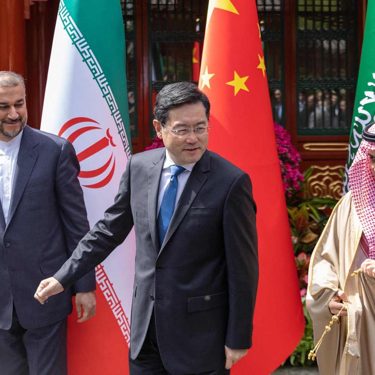 The image features three men standing together, with national flags of Iran, China, and Saudi Arabia in the background. The person on the left is wearing a suit with an Iranian flag pin, the central figure is dressed in a blue suit, and the individual on the right is wearing traditional Saudi attire. They appear to be engaged in a conversation or moment of interaction, suggesting a diplomatic or friendly meeting. The setting seems to be outdoors, possibly at a state event or formal gathering.