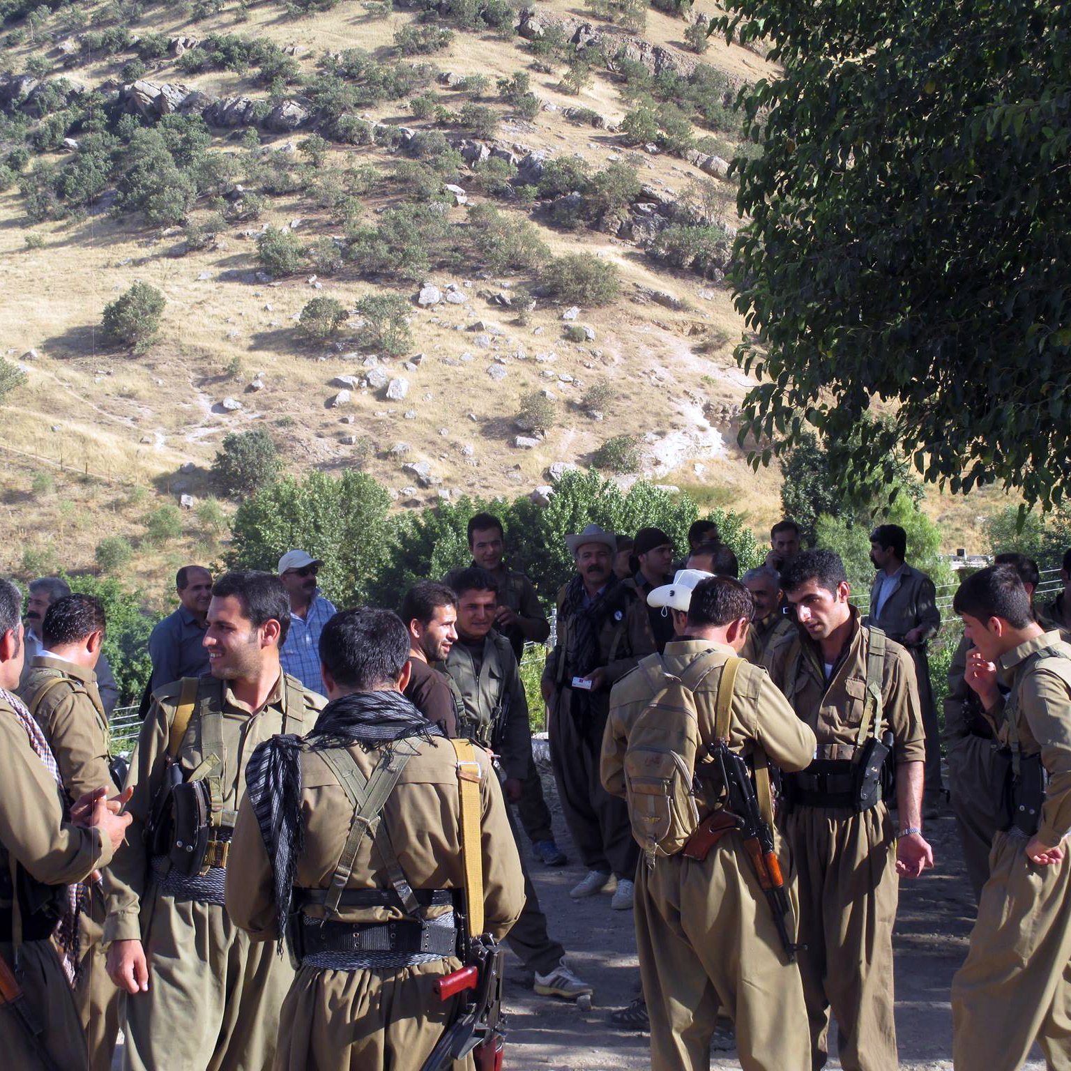 L'image montre un groupe de personnes rassemblées dans un environnement rural, avec des collines verdoyantes en arrière-plan. Les individus portent des vêtements traditionnels et certaines personnes sont armées. Ils semblent engager dans des discussions, tandis que d'autres se rassemblent autour d'eux. La scène reflète une atmosphère communautaire en milieu naturel.