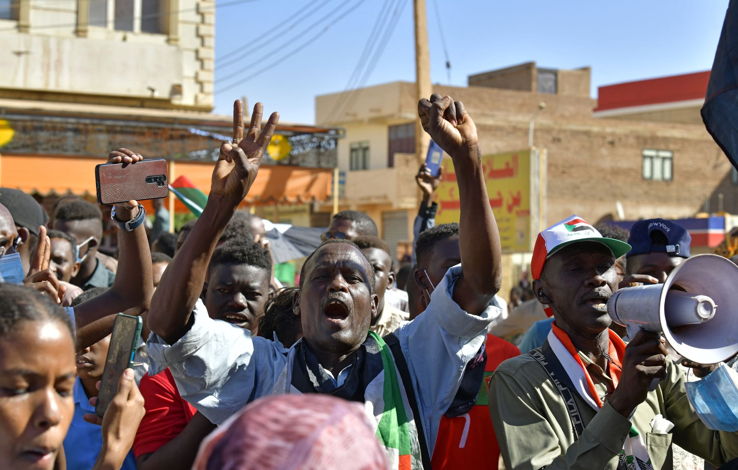 L'immagine mostra un gruppo di persone in una manifestazione vibrante. Gli individui sembrano esprimere passione e determinazione mentre sollevano le braccia e alcuni gridano slogan. Alcuni di loro indossano colori che richiamano una bandiera, suggerendo un forte attaccamento a una causa o a un movimento. È visibile anche un megafono, che indica che ci sono leader che cercano di comunicare con la folla. Lo sfondo presenta edifici e una scena urbana tipica di un contesto di protesta. L'atmosfera sembra intensa e carica di emozioni.
