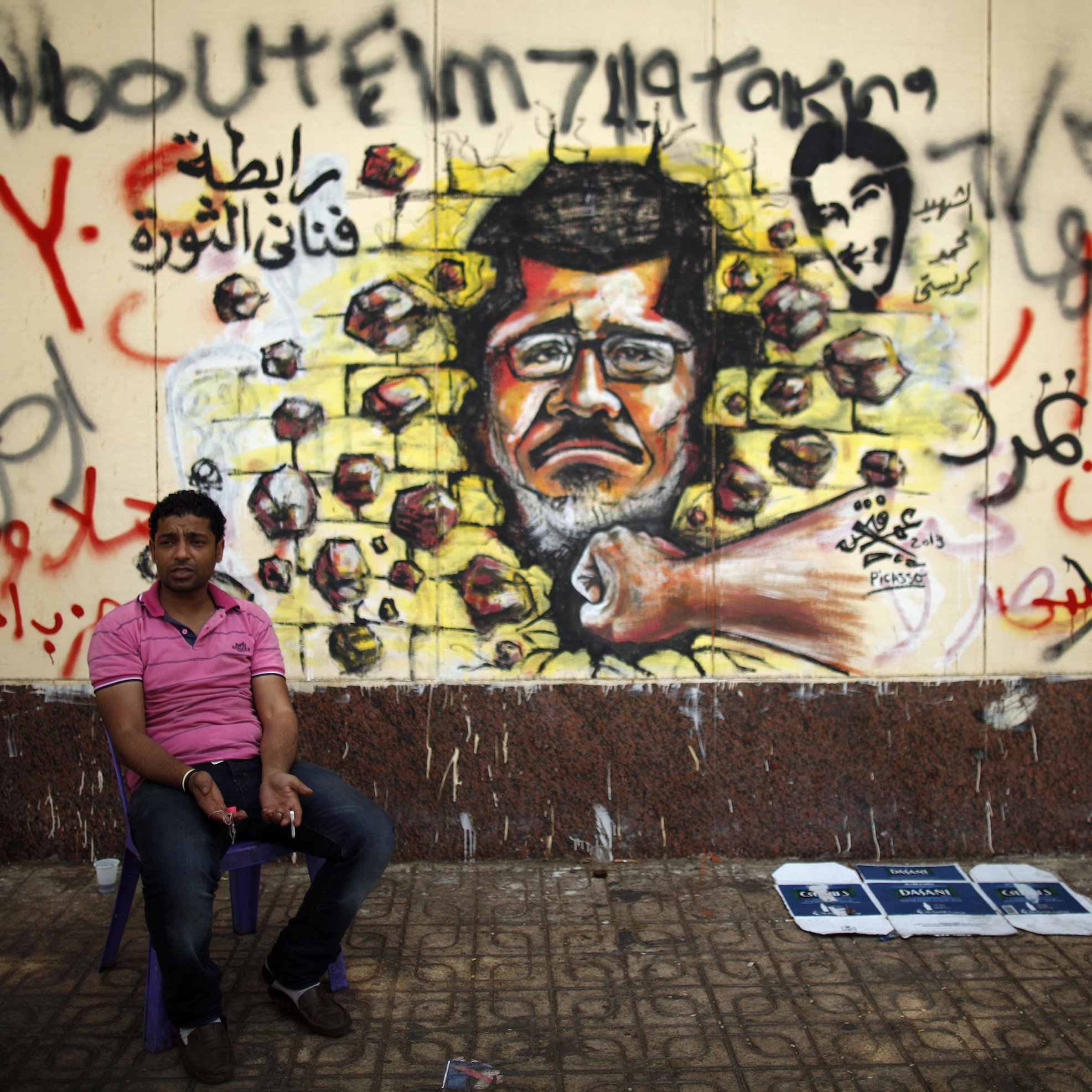 L'image montre un homme assis sur une petite chaise, devant un mur qui est recouvert d'une fresque murale. Cette fresco représente un portrait d'un homme au visage sérieux, avec des roches projetées autour de lui, symbolisant peut-être la résistance ou la lutte. Le mur est également couvert d'inscriptions en arabe et peut-être en d'autres langues, ajoutant un contexte politique ou social à la scène. L'environnement semble urbain, avec un sol en pavés et des déchets visibles à proximité.