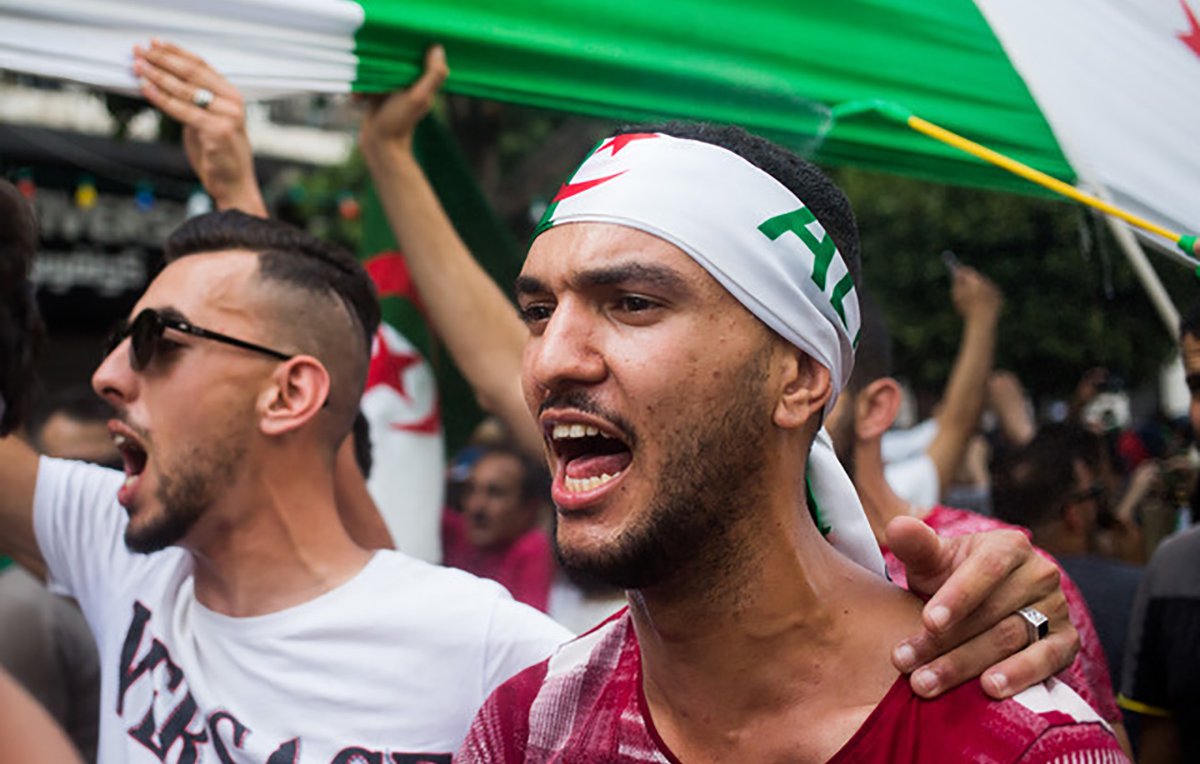 L'image montre des manifestants lors d'une protestation, probablement en lien avec une cause nationale. Deux hommes sont au premier plan : l'un porte un bandana avec des motifs évoquant l'Algérie et semble exprimer de la passion en criant. L'autre homme, à sa gauche, affiche une attitude engagée tout en portant un t-shirt à motifs. En arrière-plan, on peut voir un groupe de personnes brandissant des drapeaux, créant une ambiance de solidarité et de revendication. Les visages sont chargés d'émotion et l'atmosphère paraît énergique.