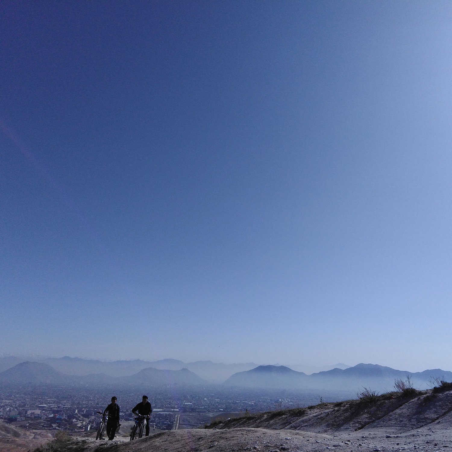 تظهر في الصورة منظر طبيعي واسع يأخذ المشاهد إلى مشهد جبلي تحت سماء زرقاء صافية. في الجزء السفلي من الصورة، يظهر شخصان يسيران على تلة مرتفعة، بينما تغطي المدينة أجزاء من السهل أدناه. تتوزع الجبال في الأفق بشكل جميل، مما يعطي إحساسًا بالاتساع والعمق. الألوان هادئة، مع وجود أعمدة خفيفة من الغيوم في السماء، مما يضفي على المشهد إحساسًا بالهدوء والسكون.