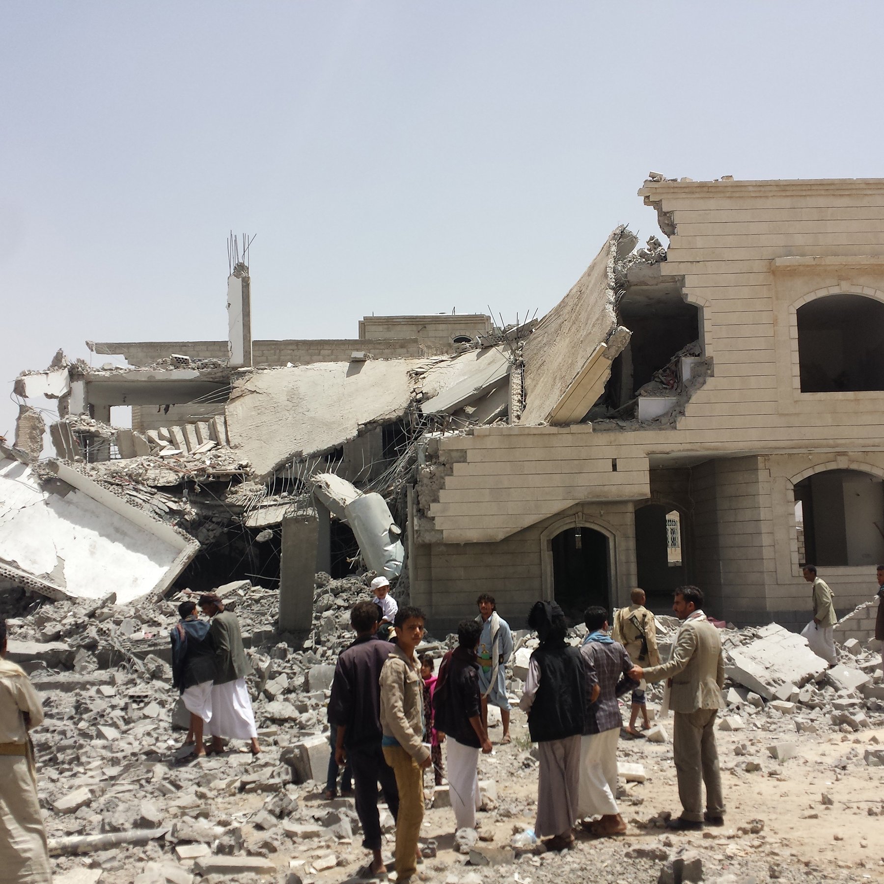 L'image montre un bâtiment partiellement détruit, avec des débris éparpillés autour. La structure semble avoir subi des dommages importants, avec un étage effondré. Des personnes se rassemblent autour des décombres, certaines observant la scène tandis que d'autres semblent discuter. Le ciel est clair et le sol est couvert de gravats. L'atmosphère semble être celle d'une enquête ou d'un recensement des dégâts.