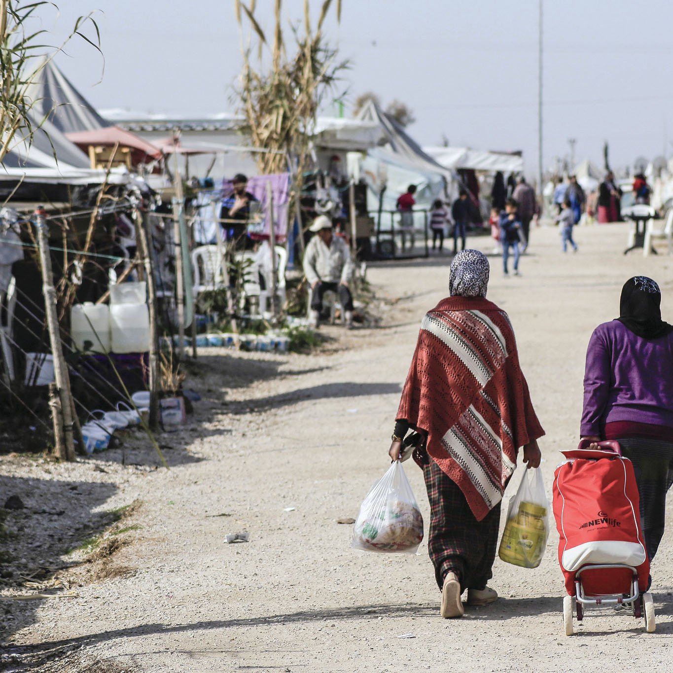 The image depicts two women walking down a dirt path in a makeshift camp. One woman carries bags of supplies, while the other pushes a stroller. On either side of the path, there are tents and structures that suggest a temporary living situation. The environment appears to be bustling with activity, as other individuals can be seen in the background, some seated or engaging in conversation. The scene conveys a sense of community within the challenges of a refugee or displaced persons camp.