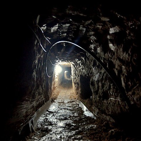 L'image montre un tunnel sombre creusé dans la terre. Les parois de la cavité sont en pierre, et le sol est inégal, probablement en terre battue. Une lumière blanche éclaire partiellement le tunnel, révélant son intérieur. Le fond du tunnel semble s'étendre à l'infini, créant une ambiance mystérieuse. Des câbles électriques sont visibles à certains endroits, indiquant qu'il pourrait y avoir des installations techniques à l'intérieur.