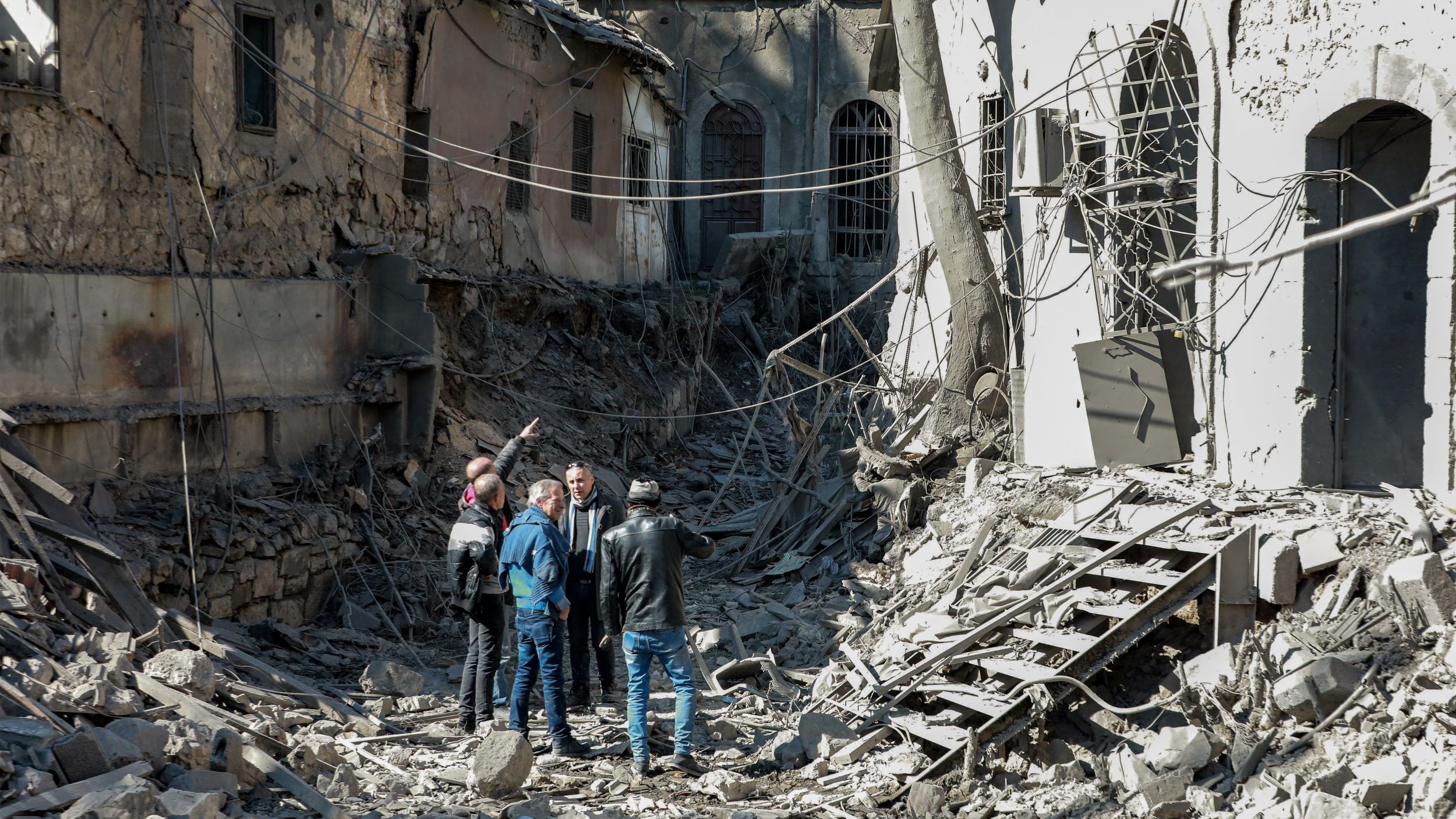L'image montre un groupe de personnes se tenant au milieu d'un paysage de destruction. Les bâtiments autour d'eux sont en ruines, avec des murs effondrés et des débris éparpillés sur le sol. Les personnes semblent discuter ou examiner les lieux, tandis que des fils électriques sont visibles dans l'environnement. L'atmosphère est marquée par un sentiment de désolation et de perte.