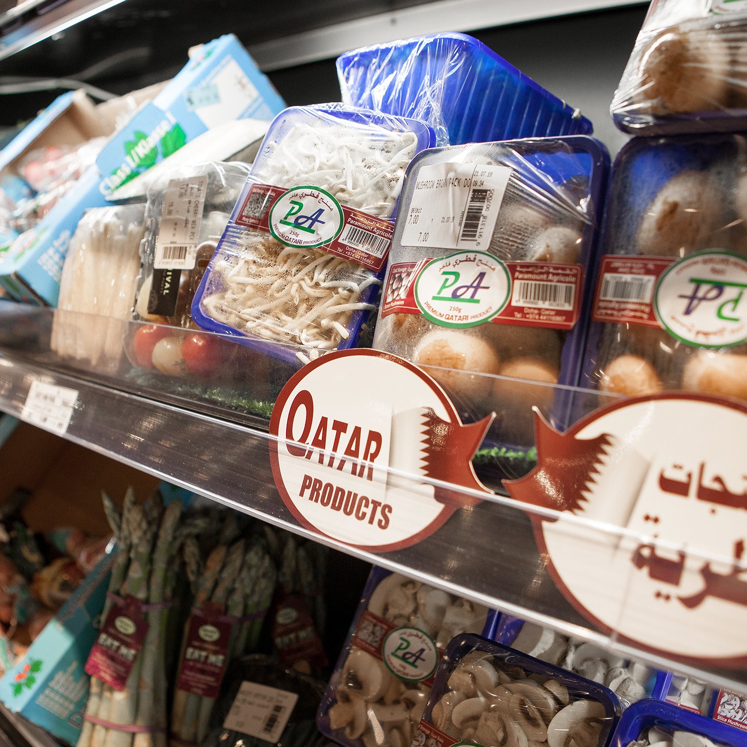 The image shows a grocery store shelf displaying a variety of packaged food items, primarily focused on fresh produce and seafood. There are containers of different types of mushrooms, noodles, and possibly other vegetables. A prominent sign labeled "Qatar Products" indicates a selection of items that are local or regional. The shelves are organized, showcasing a well-stocked array of products.