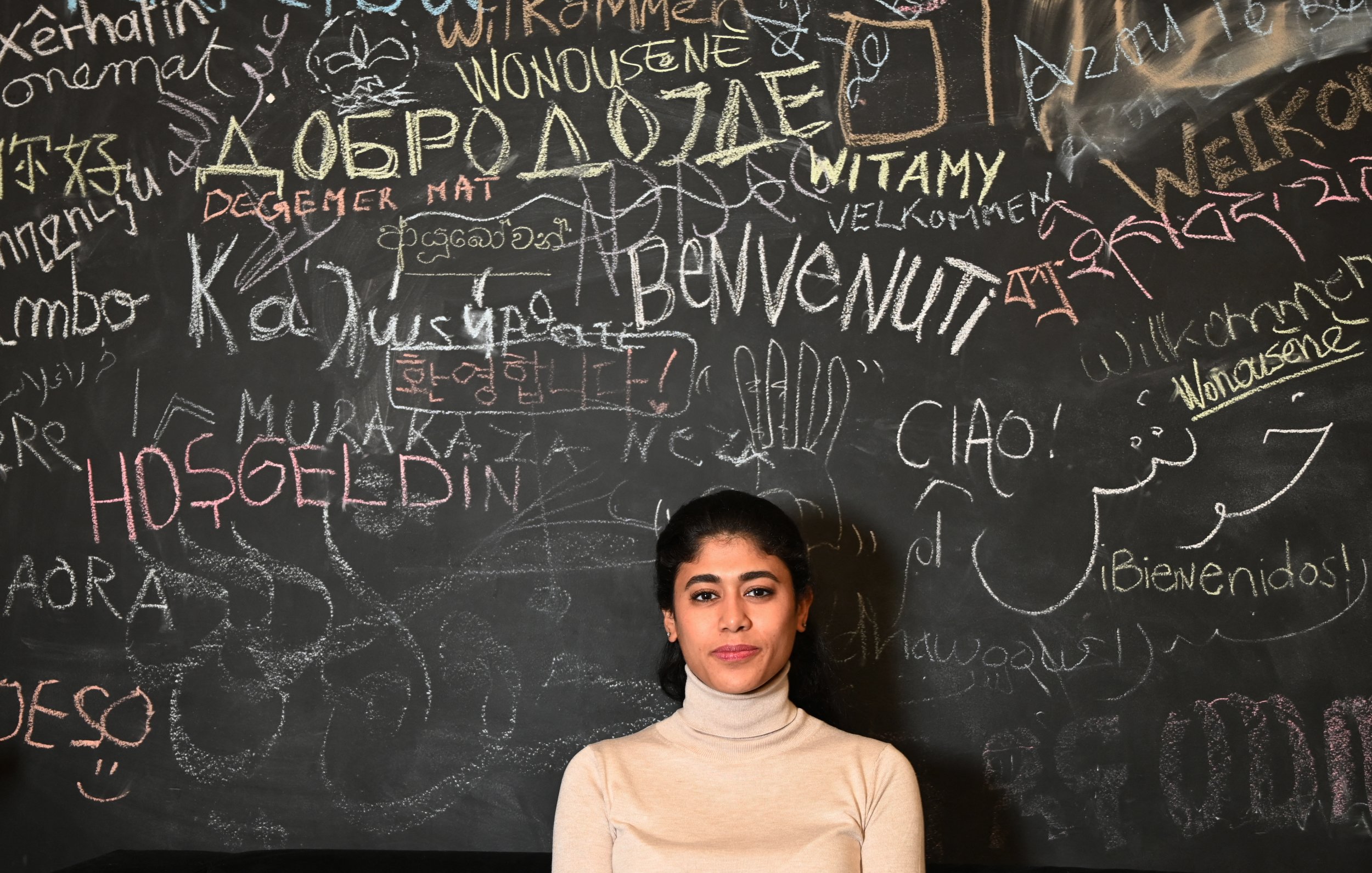 La imagen muestra a una joven posando frente a una pared de tiza oscura cubierta de múltiples palabras y frases escritas en diferentes idiomas. La mujer lleva un suéter claro y tiene el cabello recogido. La pared refleja un ambiente multicultural, con saludos y palabras de bienvenida en distintas lenguas.