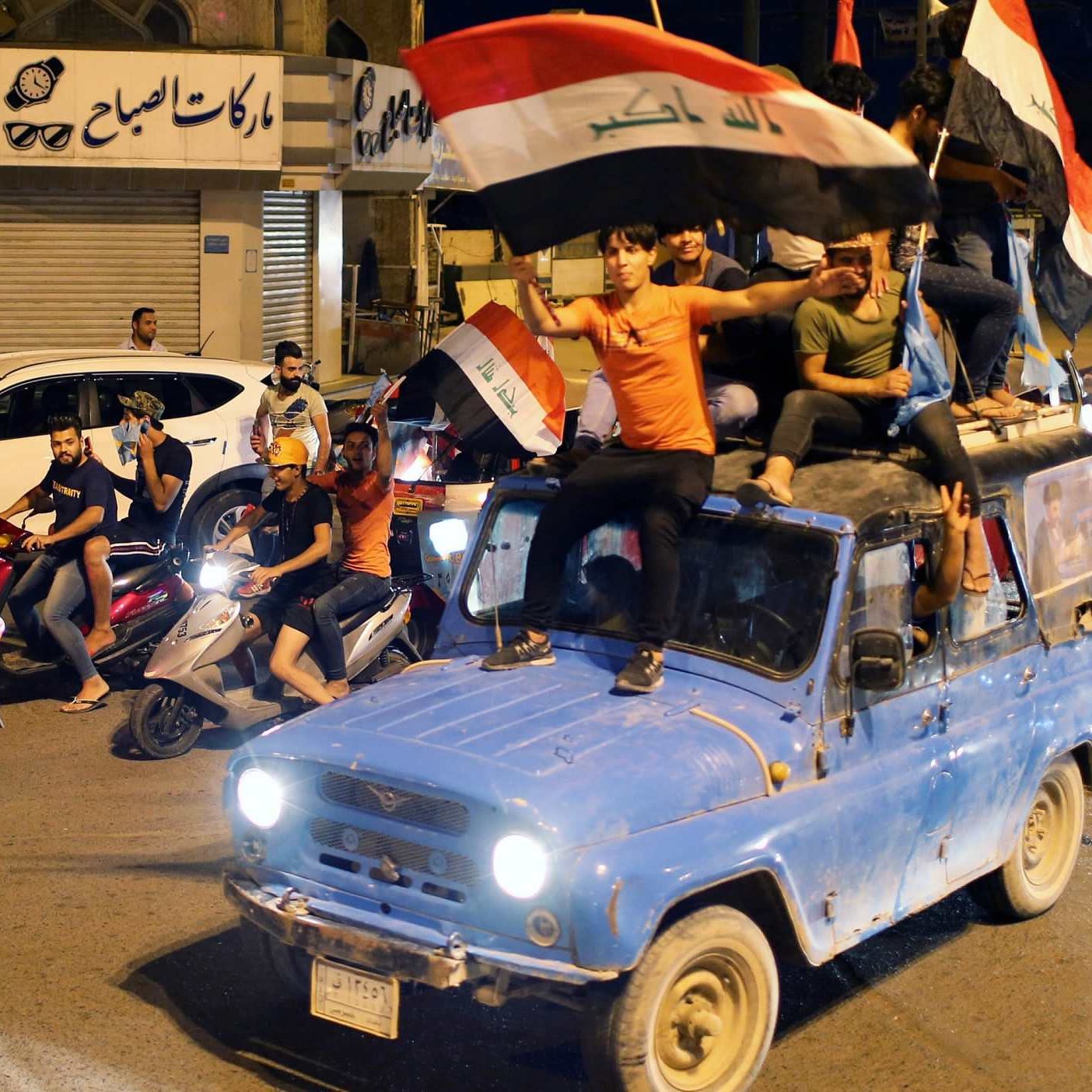 L'image représente une scène de célébration dans une rue animée. Des personnes sont rassemblées autour de véhicules, certains montent sur un ancien pickup bleu. Des drapeaux irakiens sont agités, tandis que d'autres participants conduisent des motos et des scooters. L'atmosphère est festive et dynamique, indiquant un moment de joie collective. Des bâtiments sont visibles en arrière-plan, suggérant une ambiance urbaine.