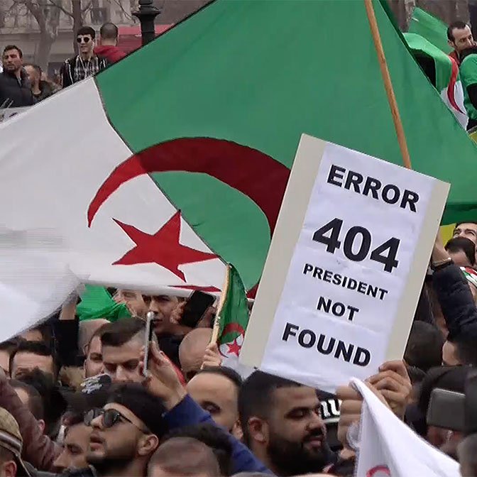 L'image montre une manifestation avec de nombreuses personnes brandissant des drapeaux algériens. Au premier plan, un manifestant tient une pancarte avec l'inscription "ERROR 404 PRESIDENT NOT FOUND", suggérant un mécontentement vis-à-vis de la situation politique. L'ambiance semble être celle d'une contestation, avec une foule réunie pour exprimer ses revendications.