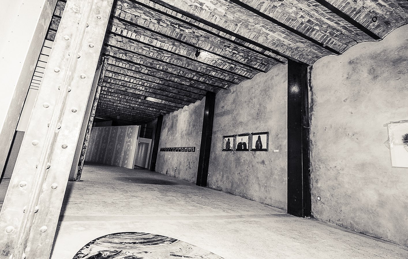 L'image présente l'intérieur d'un espace d'exposition ou d'une galerie d'art, avec de hauts plafonds en poutres de bois. Les murs sont en béton brut, créant une ambiance industrielle. Sur le mur de droite, on peut apercevoir quelques cadres accrochés, probablement contenant des œuvres d'art. Le sol est neutre, avec une texture qui semble lisse. L'éclairage met en valeur les détails architecturaux de l'intérieur, tandis que l'ensemble dégage une atmosphère contemporaine et artistique.