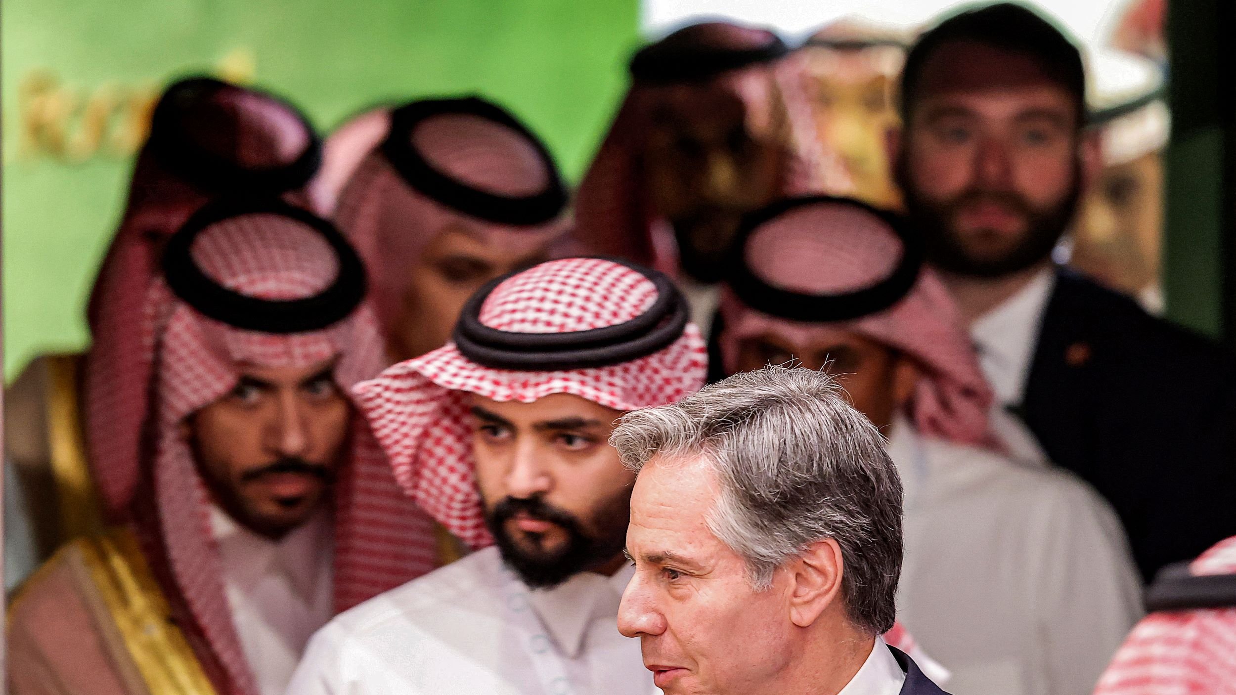 در این تصویر، تعدادی مرد که به نظر میرسد لباسهای سنتی عربی پوشیدهاند، در پسزمینهای روشن و زنده قرار دارند. در جلو، مردی با کت و کراوات به سمت راست در حال حرکت است. به نظر میرسد این تصویر مربوط به یک نشست یا مراسم رسمی باشد. فضای کلی تصویر حس تعامل و گفتوگو را منتقل میکند.