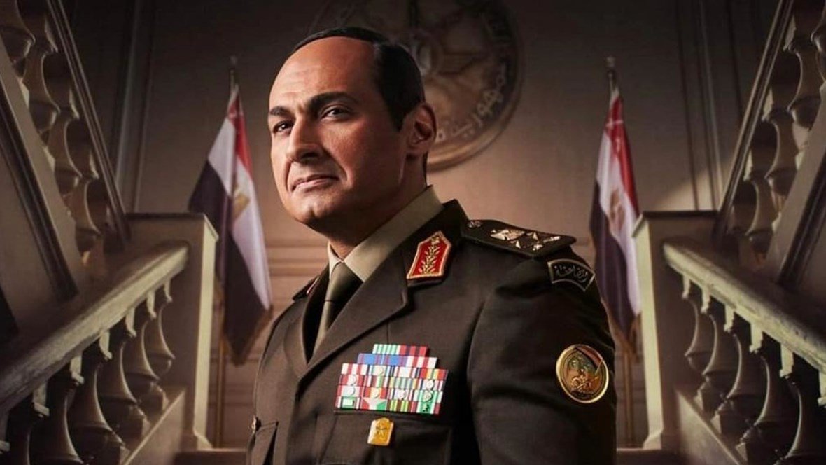 يظهر في الصورة رجل يرتدي زيًا عسكريًا يحمل العديد من الأوسمة والرتب. يبدو أنه يتخذ وضعًا رسميًا وجادًا، مع خلفية تتضمن علمين. تعكس تعابير وجهه الثقة والسلطة، كما أن التصميم العام للصورة يوحي بأجواء عسكرية أو حكومية.