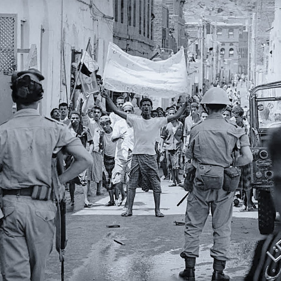 The image depicts a historical scene in a narrow street, likely during a protest or demonstration. In the foreground, a group of people is visible, with a man raising a banner or flag, indicating a spirited display. There are several individuals, including children, observing the scene. Two uniformed officers can be seen in the foreground, suggesting a presence of law enforcement. The atmosphere appears tense, with people engaged in an active moment, highlighting social or political unrest. The black-and-white format emphasizes the historical context of the image.