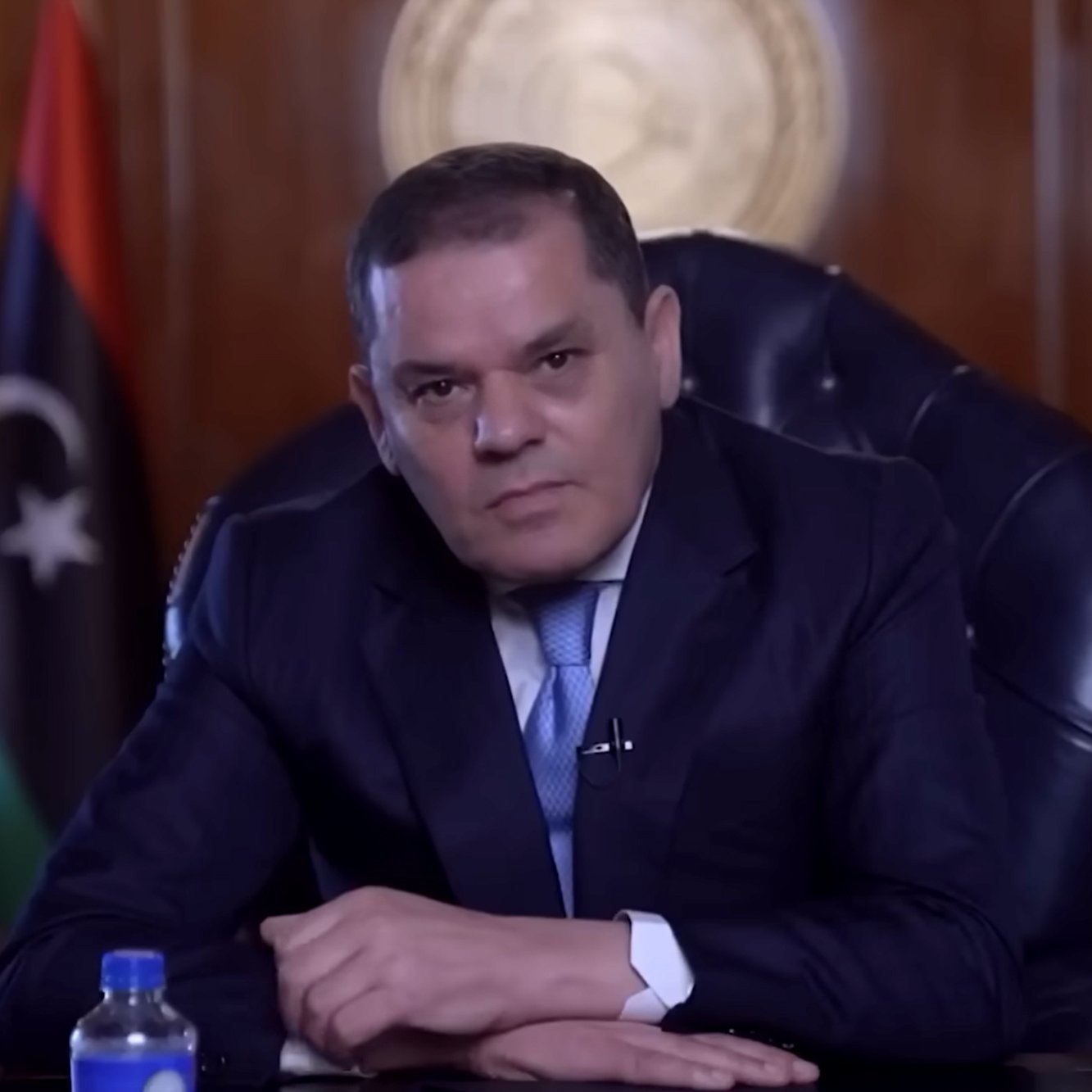 Homme en costume assis dans un bureau, drapeau libyen en arrière-plan.