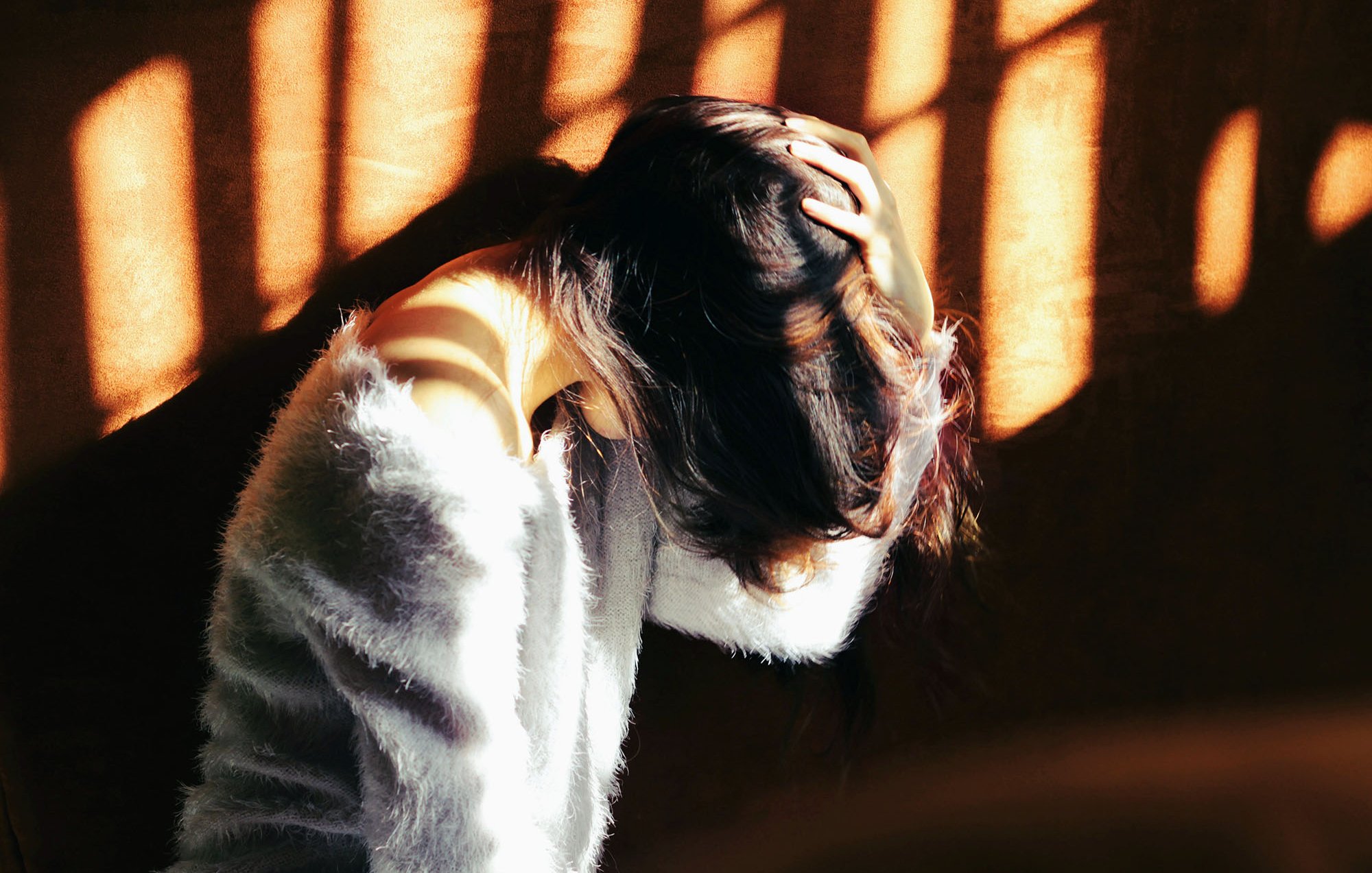 L'image montre une femme assise, penchée en avant et se tenant la tête avec ses mains. Sa posture suggère un état de réflexion ou de malaise. La lumière du soleil filtre à travers des barreaux, créant des ombres graphiques sur elle et sur le mur derrière. Elle porte un vêtement doux et pelucheux, ce qui contraste avec l'intensité de la lumière. L'ambiance générale de l'image évoque une certaine introspection ou une atmosphère mélancolique.