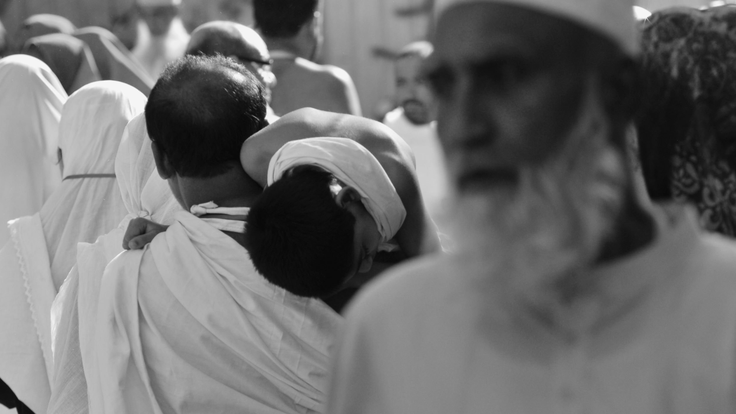 L'image montre une scène animée d'une foule, probablement lors d'un événement religieux. On peut voir un homme portant un enfant sur son dos, tandis que d'autres personnes sont visibles en arrière-plan, certaines portant des vêtements traditionnels. L'image est en noir et blanc, ce qui accentue l'atmosphère solennelle de l'événement. Les expressions des visages et les attitudes des personnes semblent refléter une ambiance de dévotion et de spiritualité.