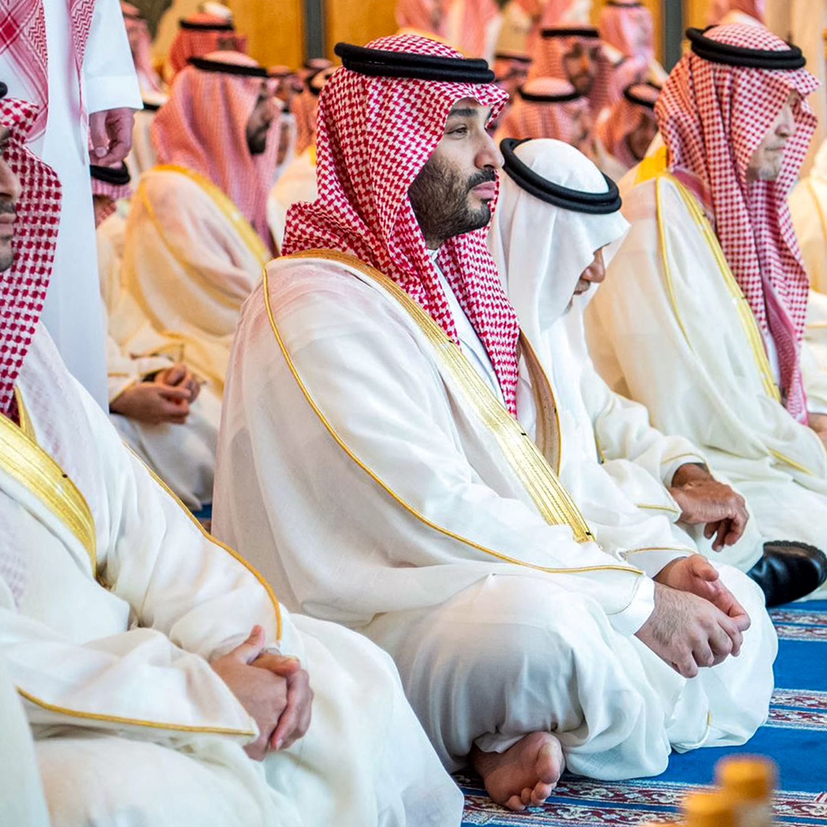 L'image montre un groupe d'hommes assis, probablement en train de prier ou de méditer. Ils portent des vêtements traditionnels saoudiens, comme des thobes blancs et des chapeaux appelés ghutras, souvent ornés de bandes rouges et blanches. L'atmosphère semble empreinte de sérénité et de respect, reflet d'un moment spirituel partagé. Le décor autour d'eux est élégant et peut inclure des éléments architecturaux typiques d'un lieu de culte.
