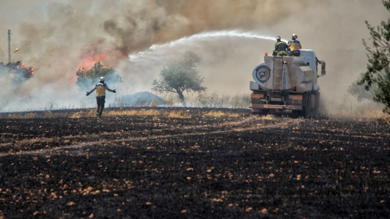 L'image montre une scène de lutte contre un incendie. On voit des pompiers en action, utilisant un camion-citerne pour éteindre les flammes qui s'élèvent dans la fumée. Au premier plan, un pompier semble se diriger vers le feu, tandis que l'arrière-plan montre un paysage noirci, témoignant des dégâts causés par l'incendie. Les arbres sont entourés de fumée, créant une atmosphère de tension et d'urgence.