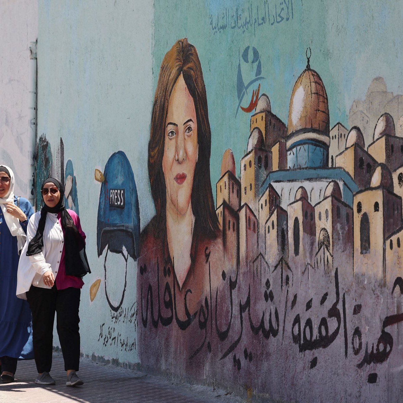 L'immagine mostra due donne che camminano lungo una strada, accanto a un murale. Il murale ritrae una donna con un cappello alla "press" e un volto espressivo. Sullo sfondo, si possono vedere edifici caratteristici, e ci sono scritte in arabo che possono fare riferimento a temi di libertà di stampa o giustizia. La scena è vivace e riflette una cultura ricca e una narrazione importante legata ai diritti dei giornalisti.