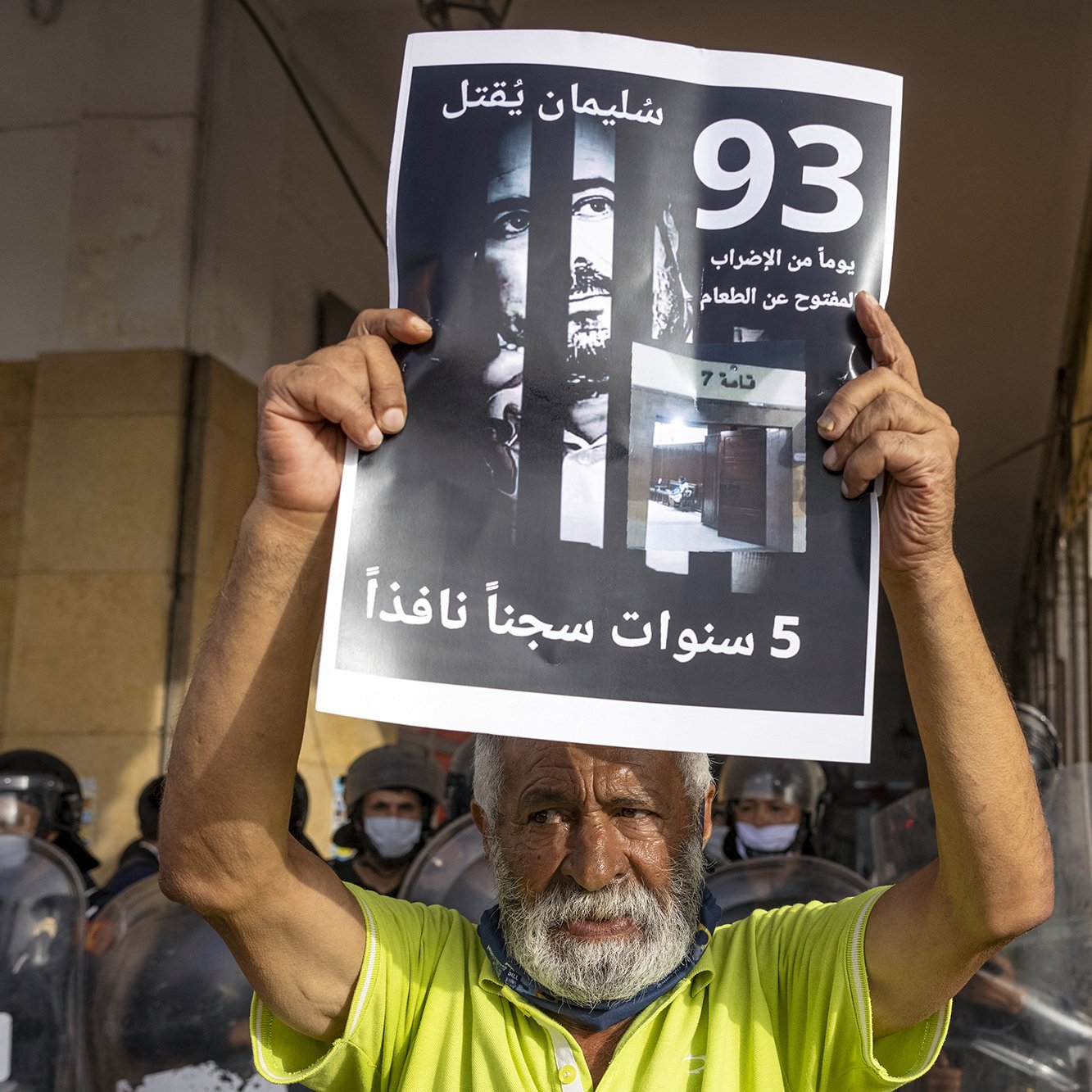 L'immagine mostra un uomo anziano che tiene in alto un cartello. Sul cartello si possono vedere scritte in arabo e un'immagine di volti, che suggerisce un tema di protesta o di richiesta di giustizia. Sullo sfondo ci sono agenti di polizia in uniforme antisommossa, suggerendo che l'evento si svolge in un contesto di tensione. L'anziano sembra determinato mentre partecipa a questa manifestazione.