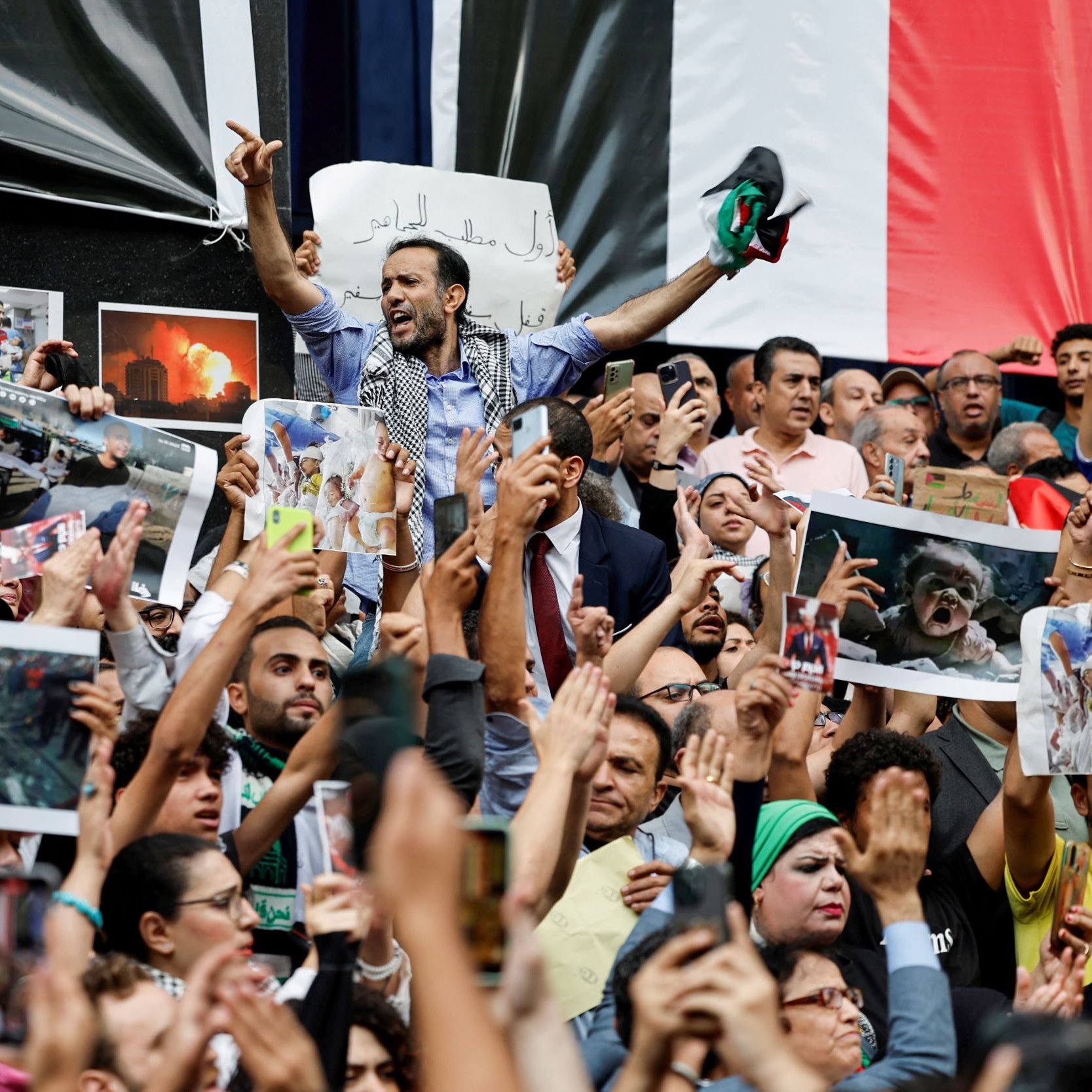 این تصویر یک تجمع بزرگ را نشان می‌دهد که مردم در آن با شور و هیجان حضور دارند. تعدادی از افراد در حال نشان دادن تصاویر و پلاکاردهایی هستند. برخی از معترضان در حال فریاد زدن یا ابراز احساسات خود هستند و مشخص است که موضوعی برای آنها اهمیت دارد. در پس‌زمینه، پرچم‌ها و تصاویری از وقایع یا شخصیت‌های مهم ممکن است مشاهده شود. جو کلی تصویر پر از انرژی و احساسات قوی است.