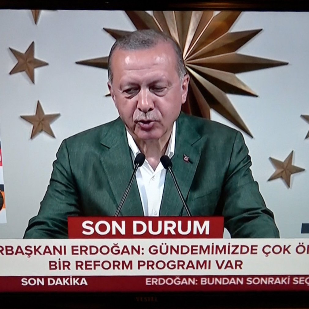 L'image montre un homme en train de prendre la parole lors d'une conférence, probablement un discours politique. Il porte une veste verte et se tient devant un grand fond décoré, avec ce qui semble être un symbole national ou institutionnel. En bas de l'écran, il y a un bandeau rouge qui indique "SON DURUM" et mentionne "CUMHURBAŞKANI ERDOĞAN" avec un message sur des réformes importantes. Sur le côté gauche, des informations sur des élections et des résultats sont affichées, suggérant que l'événement est lié à des élections en Turquie.