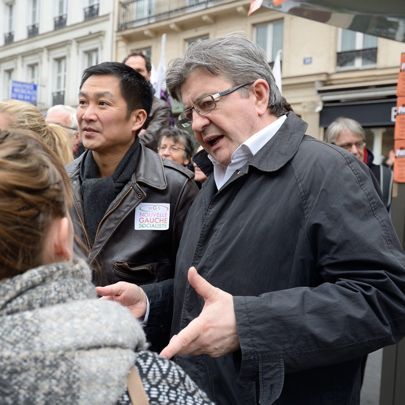 L'image montre un groupe de personnes en train de discuter dans une rue, probablement lors d'un événement public. On peut voir deux hommes au premier plan, l'un d'eux portant une veste sombre. Ils semblent engagés dans une conversation avec des femmes. L'ambiance semble dynamique et interactive, suggérant un échange d'idées ou de points de vue. Les bâtiments en arrière-plan indiquent qu'ils se trouvent dans un environnement urbain.