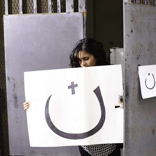 L'image montre une femme se tenant devant une porte grise. Elle tient une grande affiche présentant un symbole souriant, accompagnée d'un plus et d'un autre symbole. À côté, il y a une petite affiche accrochée à la porte. L'ensemble de la scène évoque une forme d'expression artistique, mélangeant des éléments de surprise et de réflexion.