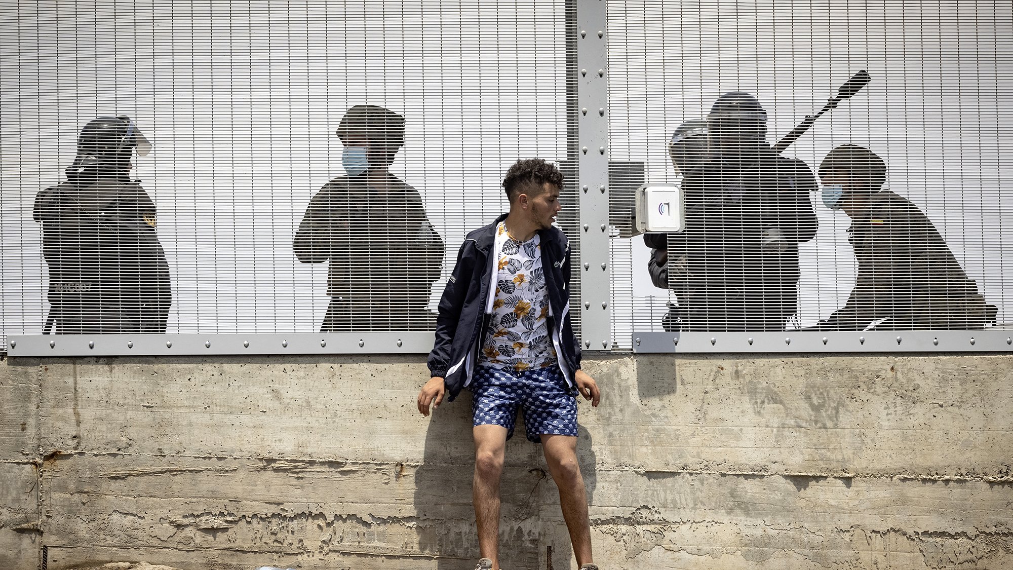 L'image montre un jeune homme debout contre un mur, habillé d'un haut sombre et d'un short à motifs. Il semble être en train de faire face à une situation tendue. Derrière lui, à travers une clôture en métal, on aperçoit des silhouettes de personnes en uniforme, apparemment des agents de la sécurité, qui semblent se préparer à intervenir. L'atmosphère est intense et suggère un contexte de conflit ou de protestation. Les ombres des agents renforcent le sentiment de pression ou de confrontation.