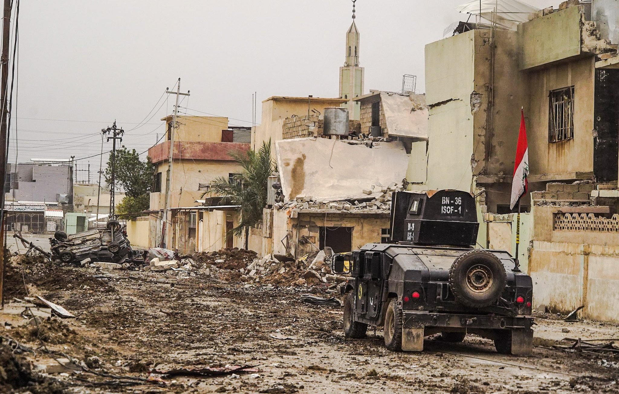 الصورة تظهر مشهداً من منطقة متضررة بسبب نزاع أو حرب. هناك الكثير من الخراب في المباني المحيطة، حيث تبدو العديد منها مدمرة أو شبه مدمرة. في منتصف الصورة، توجد مركبة عسكرية، يبدو أنها تُستخدم في عمليات الأمن أو الاستجابة الطارئة. تلوح في الأفق مئذنة مسجد، مما يشير إلى وجود مجتمع محلي. الشوارع تبدو مليئة بالتراب والحطام، مما يعكس آثار الدمار الذي لحق بالمنطقة.