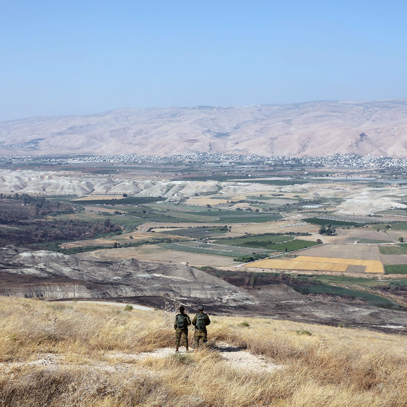 L'image montre un paysage montagneux et rural. Au premier plan, deux personnes se tiennent de dos, probablement des soldats ou des randonneurs, observant la vallée en contrebas. La vallée est composée de champs et de terrains cultivés, avec des couleurs variées allant du vert au jaune. À l'arrière-plan, des collines et des montagnes se profilent sous un ciel bleu clair. L'ensemble dégage une impression de tranquillité et de vaste espace naturel.