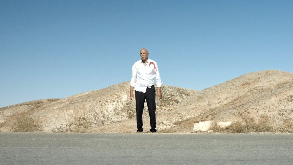 L'image montre un homme debout au milieu d'une route désertique, avec des collines arides en arrière-plan. Il porte une chemise blanche qui semble tâchée de sang et un pantalon noir. Le ciel est clair et bleu, ce qui souligne l'ambiance isolée et dramatique de la scène. Les éléments autour de lui donnent une impression de solitude et de tension.