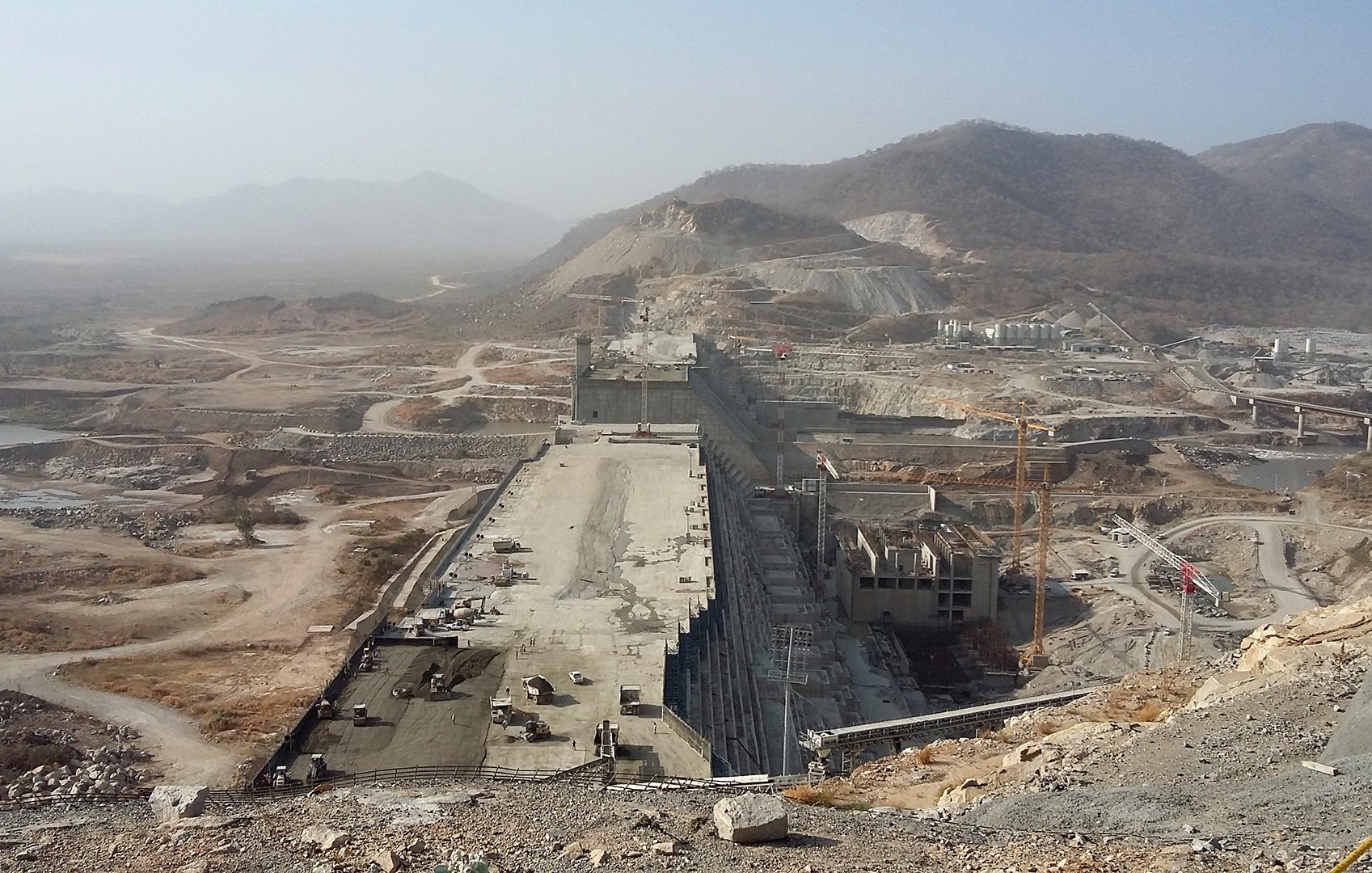 L'image montre un grand chantier de construction, probablement d'un barrage ou d'une infrastructure hydraulique. On peut voir de vastes terrassements, des routes en cours de construction et des engins lourds. L'environnement est aride, avec des collines et des montagnes à l'arrière-plan. Le ciel est légèrement brumeux, ce qui donne une atmosphère poussiéreuse à la scène. Généralement, l'image représente l'ampleur d'un projet d'ingénierie majeur.