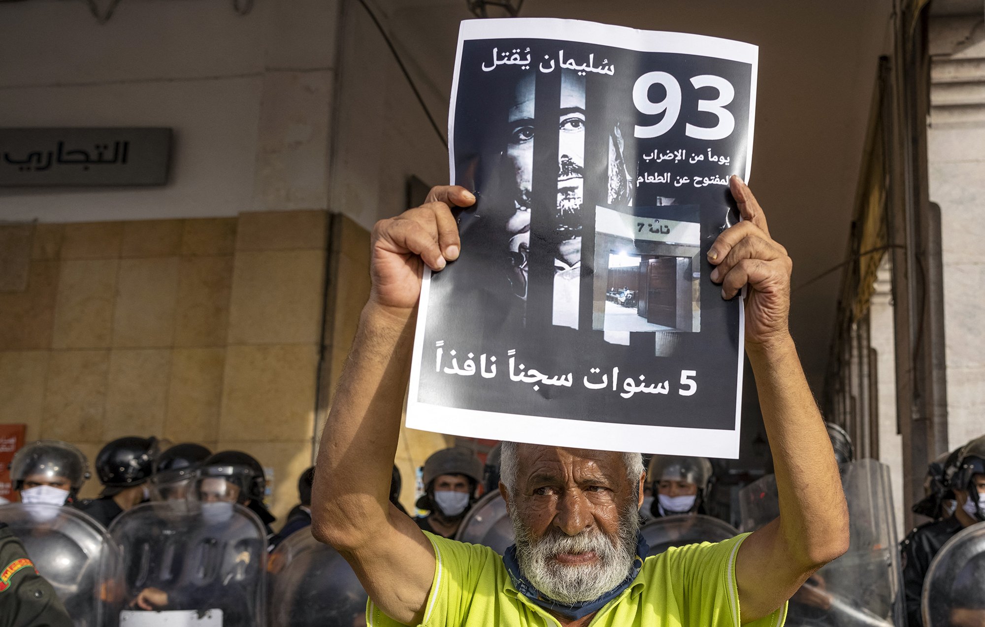 L'immagine mostra un uomo anziano che tiene in alto un cartello. Sul cartello si possono vedere scritte in arabo e un'immagine di volti, che suggerisce un tema di protesta o di richiesta di giustizia. Sullo sfondo ci sono agenti di polizia in uniforme antisommossa, suggerendo che l'evento si svolge in un contesto di tensione. L'anziano sembra determinato mentre partecipa a questa manifestazione.