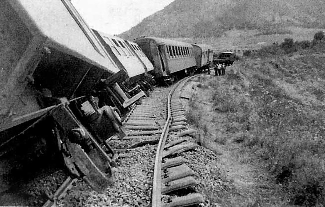 L'image montre un accident de train. Plusieurs wagons sont déraillés, penchés sur le côté, tandis que les rails semblent déformés. En arrière-plan, on aperçoit des montagnes et des personnes qui semblent inspecter les lieux. L'atmosphère est celle d'un événement tragique, évoquant une situation d'urgence.
