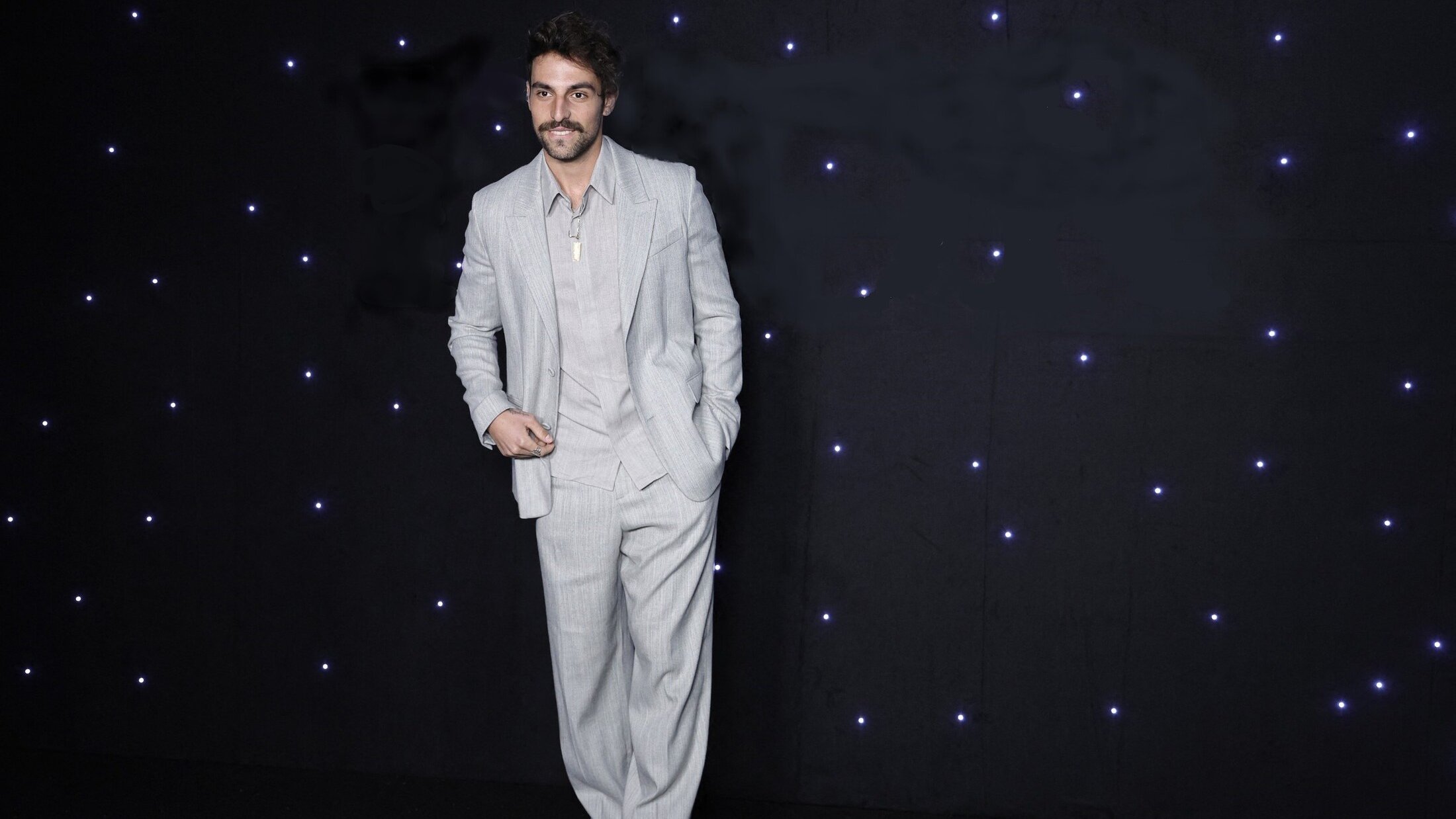 L'immagine mostra un uomo in piedi su uno sfondo scuro, illuminato da piccole luci bianche che brillano. Indossa un completo grigio chiaro, caratterizzato da pantaloni ampi e una giacca ben tagliata. La sua camicia è di un tono chiaro e ha il colletto aperto. L'uomo ha un sorriso e porta dei baffi, dando un'aria elegante e sicura di sé.