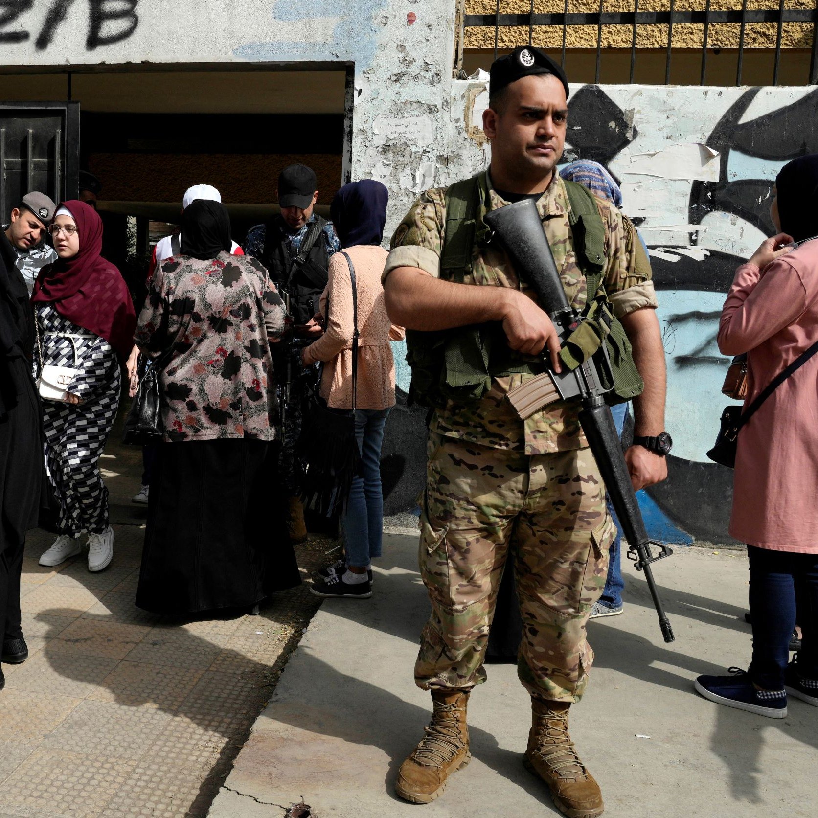 تظهر في الصورة مجموعة من الأشخاص يتجمعون في مكان يبدو أنه مدخل لمبنى، حيث يقف جندي مسلح أمام المدخل. يرتدي الجندي زيًا عسكريًا ويحمل سلاحًا، بينما تتنوع ملابس الأشخاص الذين حوله، حيث ترتدي بعض النساء العباءات والحجاب. تعكس الصورة أجواء من الحذر والاحتياط، مع وجود جندي للحماية أو تنظيم الصفوف.
