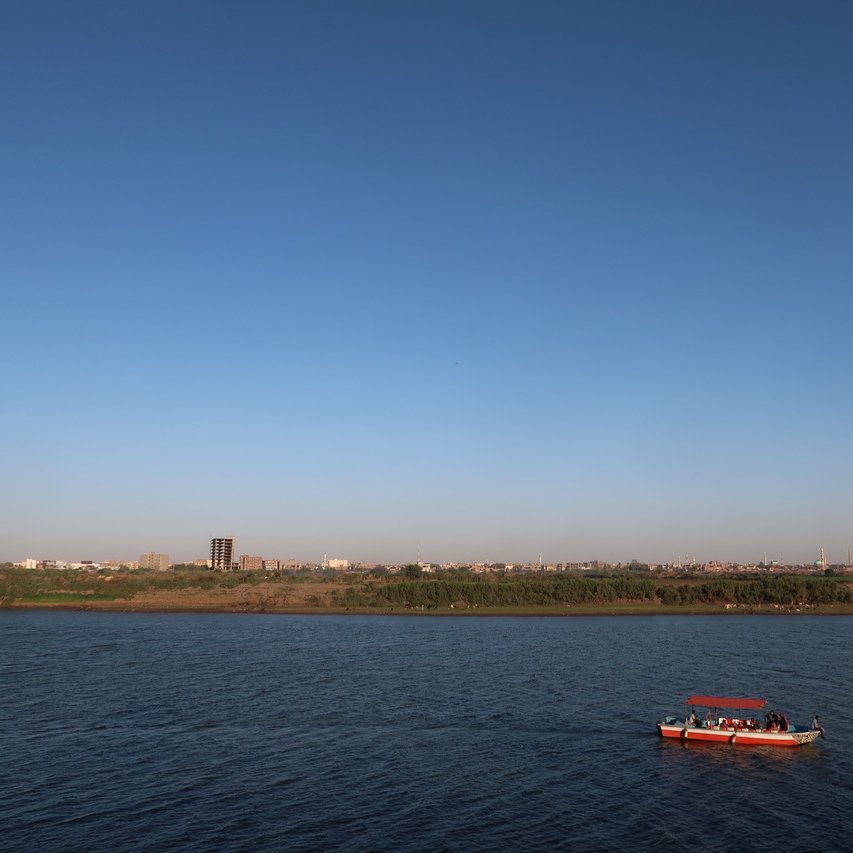 The image depicts a serene river landscape under a clear blue sky. In the foreground, a small boat with a red canopy is floating on the water, which reflects the sky. In the background, there is a sandy shore lined with greenery, and a distant cityscape can be seen, with buildings rising against the horizon. The overall scene conveys a peaceful and picturesque atmosphere.