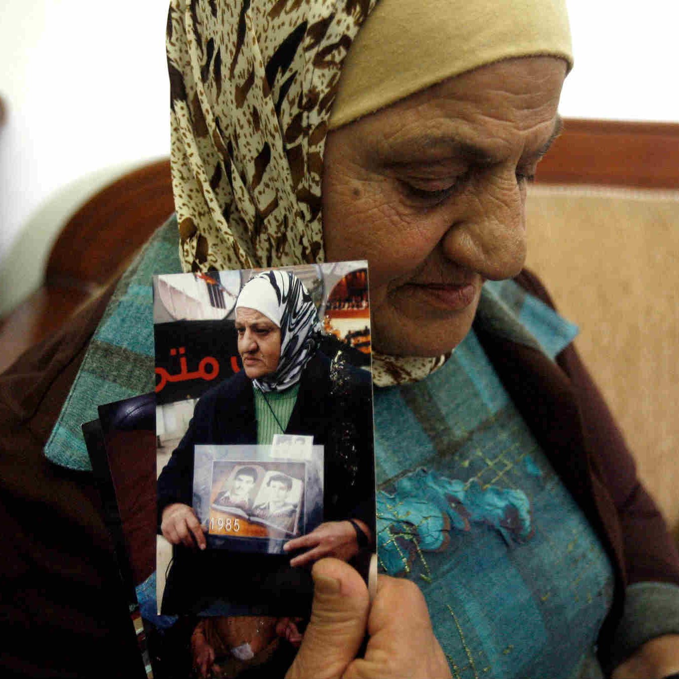 L'image montre une femme âgée portant un foulard, assise sur un canapé. Elle tient des photos en noir et blanc dans ses mains. Son expression semble empreinte de nostalgie ou de tristesse, indiquant peut-être qu'elle évoque des souvenirs douloureux. L'environnement semble simple, avec des murs clairs et un décor sobre en arrière-plan. La scène transmet une forte charge émotionnelle, suggérant un lien personnel avec les images qu'elle tient.