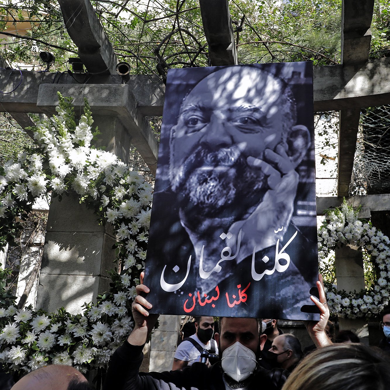 L'immagine mostra una persona che tiene in alto un grande cartello. Il cartello presenta un ritratto in bianco e nero di un uomo con una barba, accompagnato da un testo in arabo che significa "Siamo tutti Libano". Sullo sfondo, ci sono decorazioni floreali e un ambiente che sembra essere un luogo commemorativo. La scena trasmette un senso di unità e solidarietà.