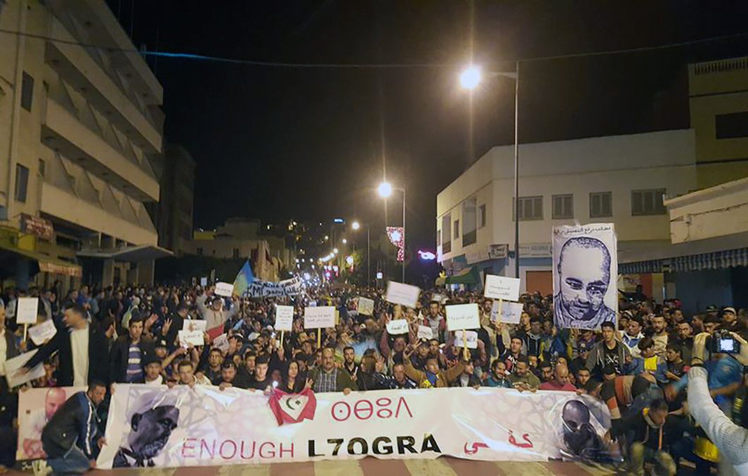 The image depicts a large crowd gathered in a street at night, participating in a protest or demonstration. Many individuals hold signs, and a prominent banner at the front reads "ENOUGH L7OGRA." There is also a large portrait of a person displayed, likely related to the cause of the protest. The atmosphere appears charged, with people expressing their views collectively in a public space. Streetlights illuminate the scene, highlighting the participants and their messages.